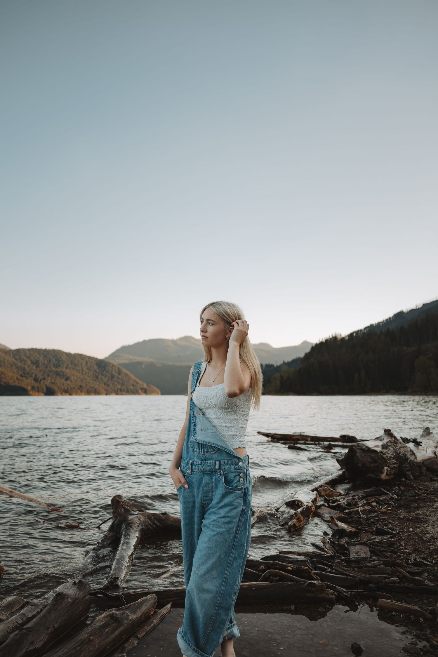 Young woman in denim overalls standing on driftwood by a serene mountain lake at dusk.