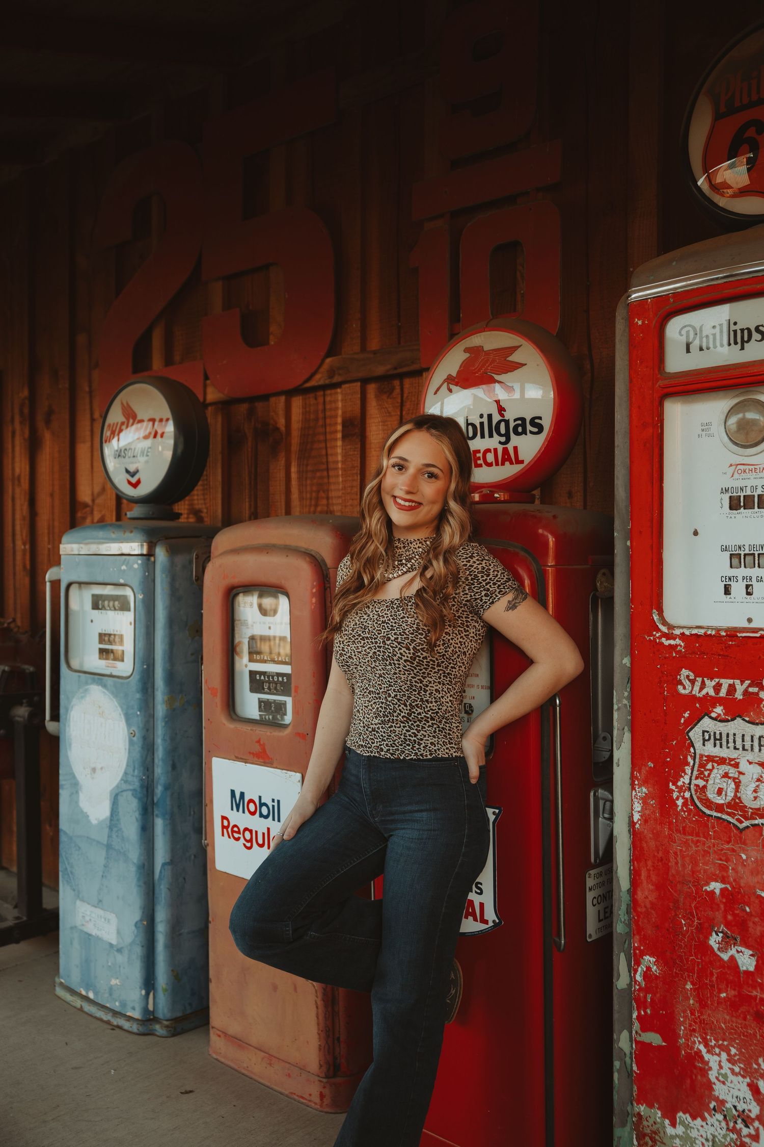 Young woman in leopard top and jeans posing by vintage Phillips 66 and Mobilgas gas pumps at a retro station.