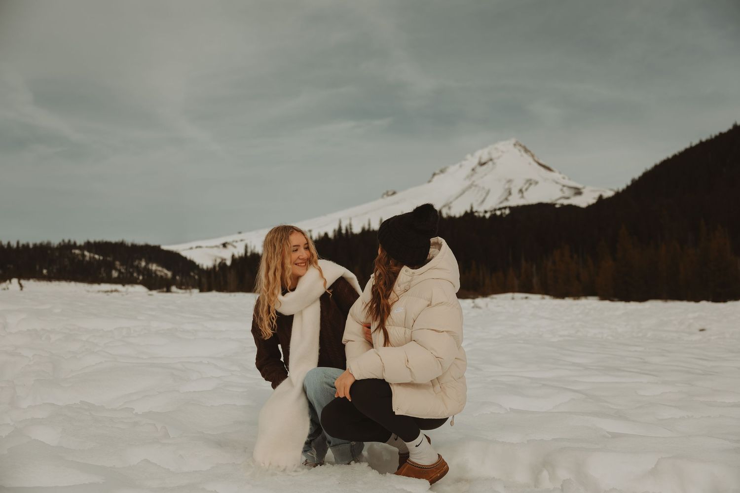 Two women laughing together in a snowy mountain landscape wearing winter coats.