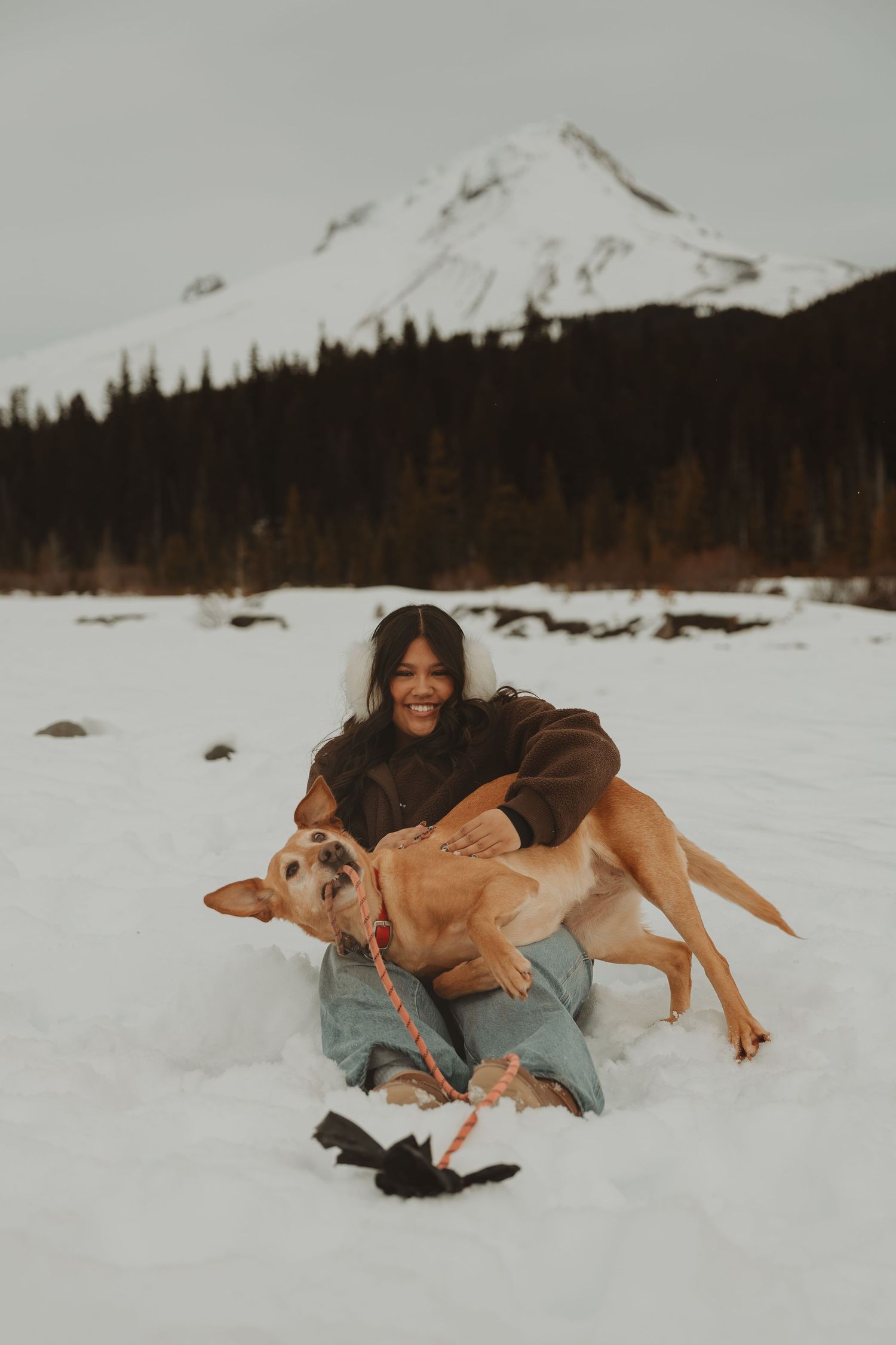 Laughing woman plays with golden dog in snow with snow-capped mountain visible in background.