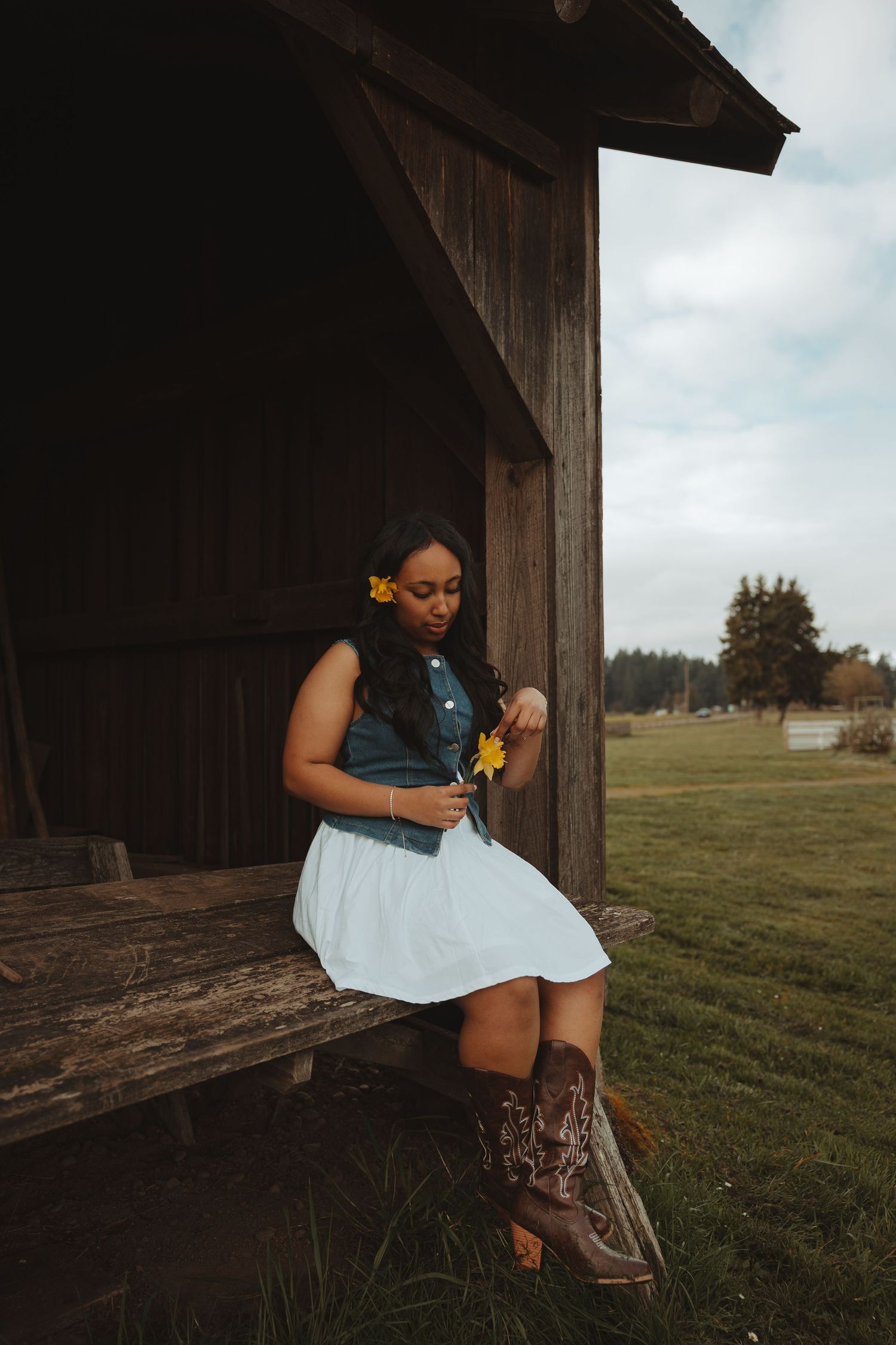 A person in a white dress and denim jacket sits on a rustic wooden structure in a rural field setting.