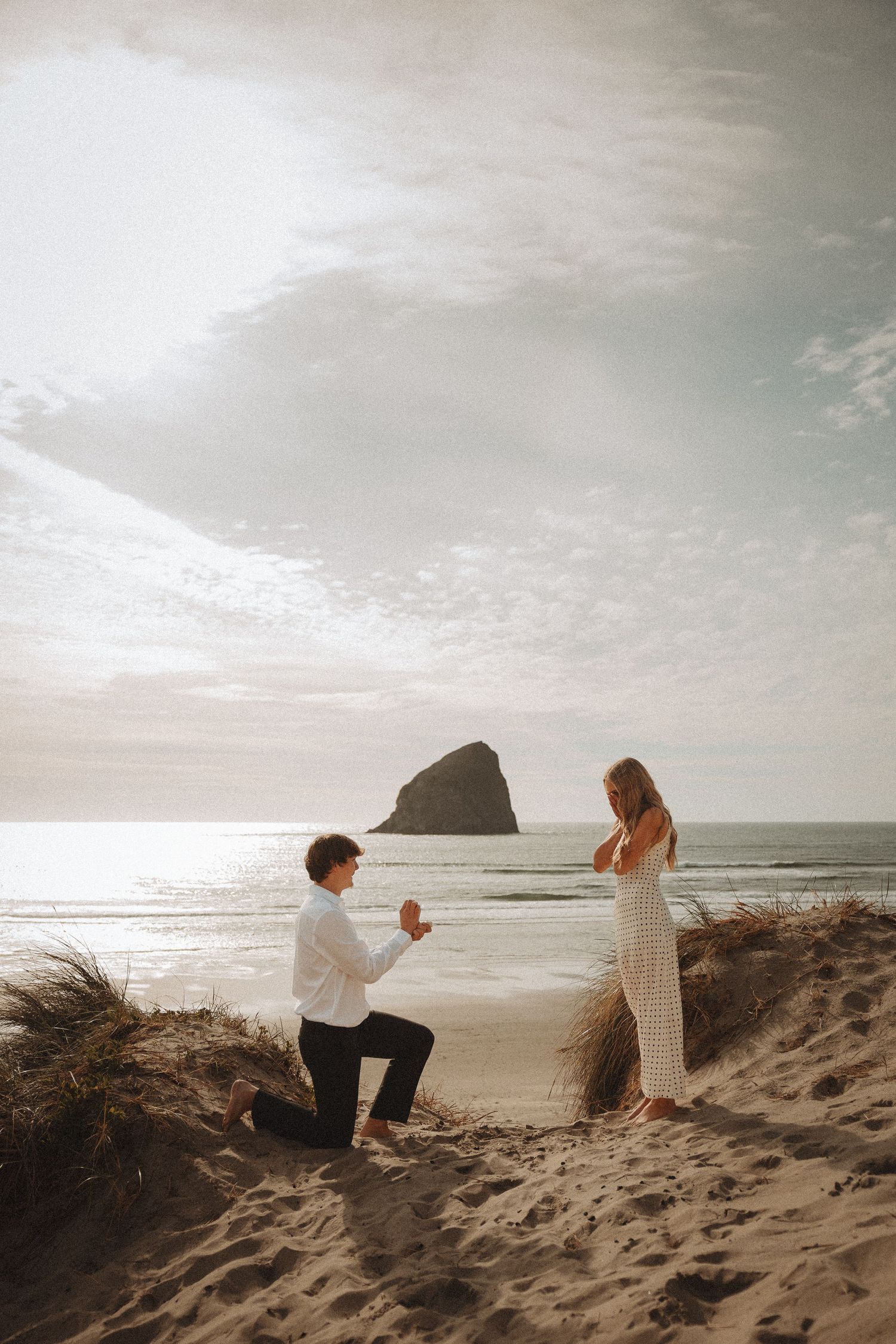 Man proposes to woman on sandy beach at sunset with dramatic rock formation in background.