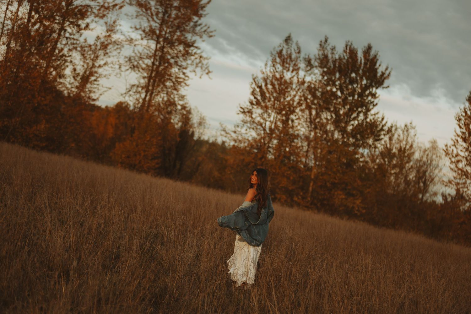 People walking through a tall grass field at sunset with trees silhouetted in the background.