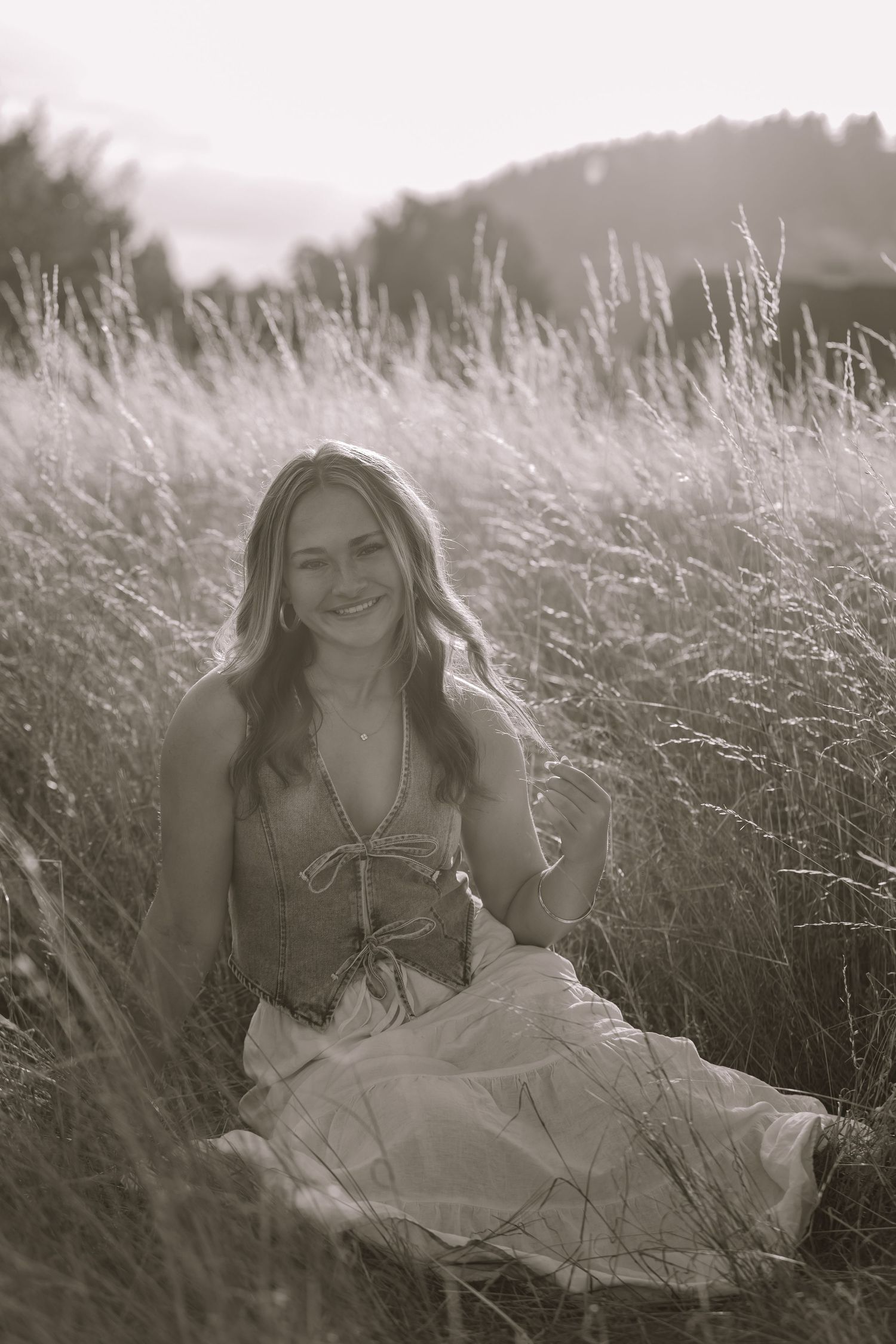 A person in a white dress lies in a sunlit field of tall grass creating a dreamy black and white portrait series.