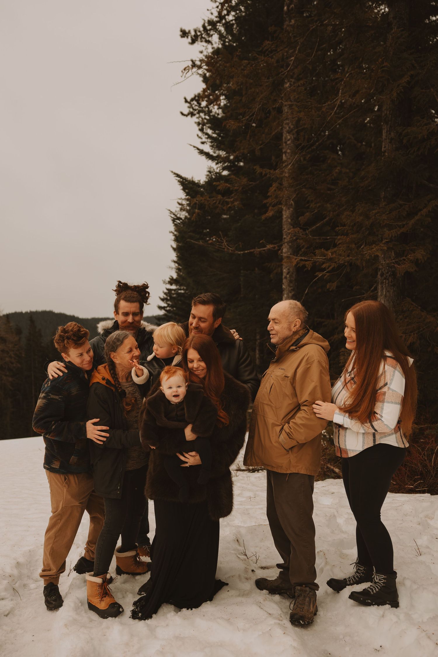 A group wearing warm winter clothes plays together in the snow against a backdrop of tall evergreen trees at sunset.
