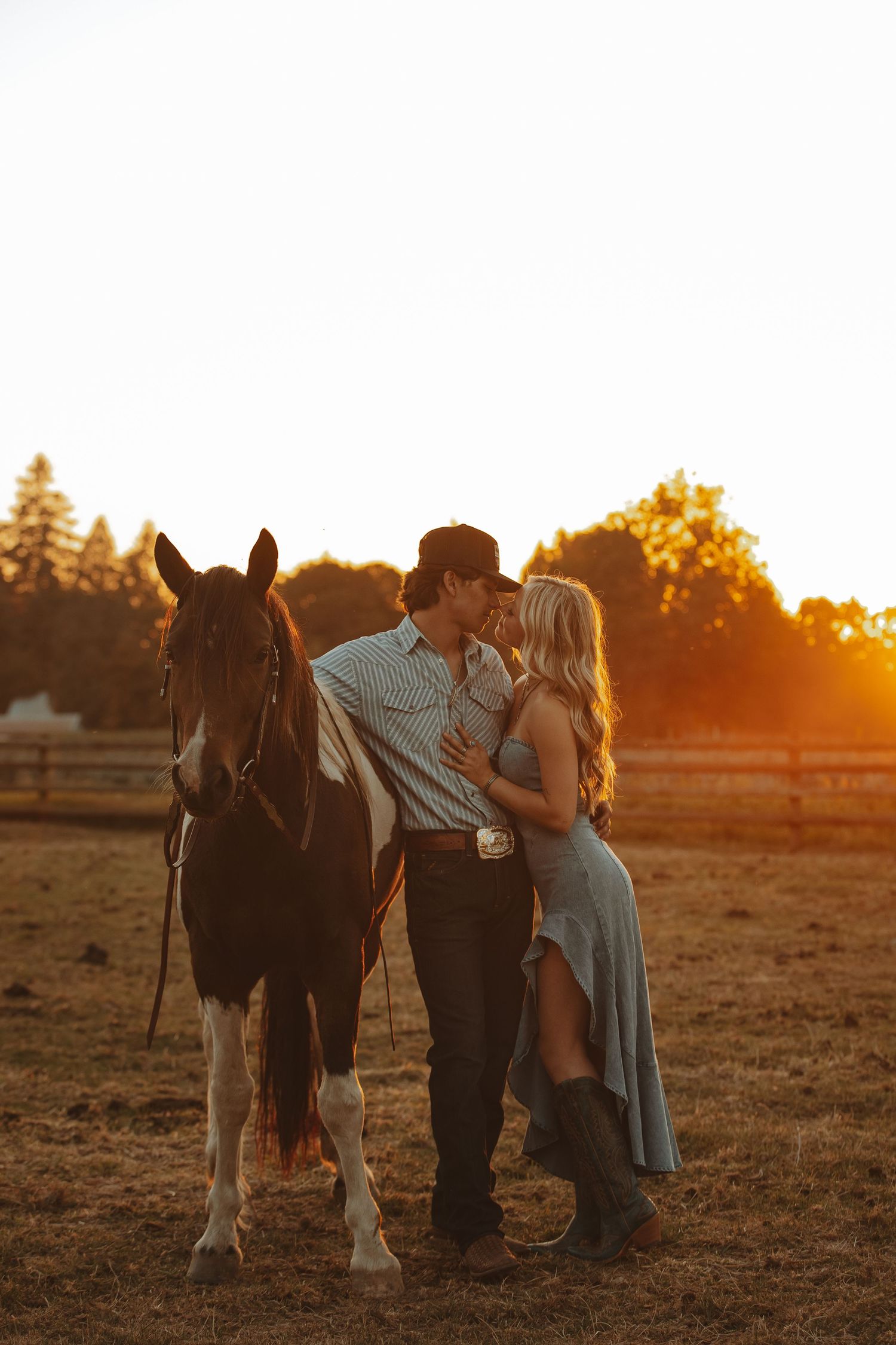 Couple kisses beside a horse during golden sunset on ranch.