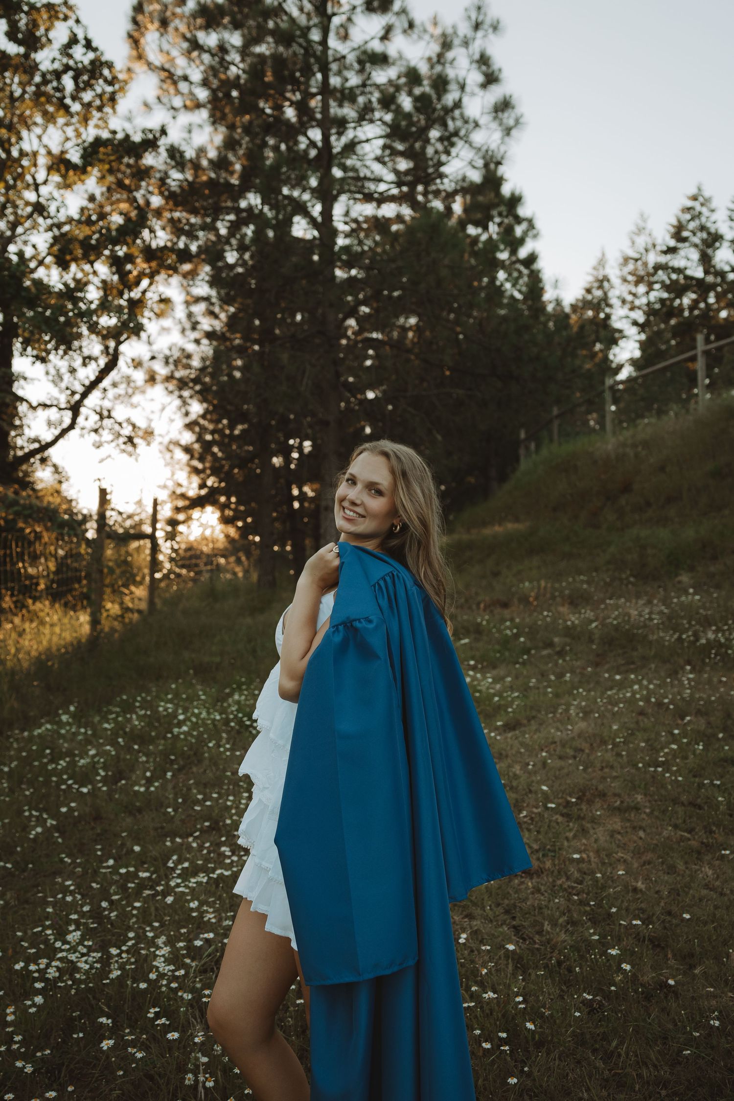 Smiling blonde woman in white dress holding blue graduation gown in golden hour meadow with wildflowers and pine trees.