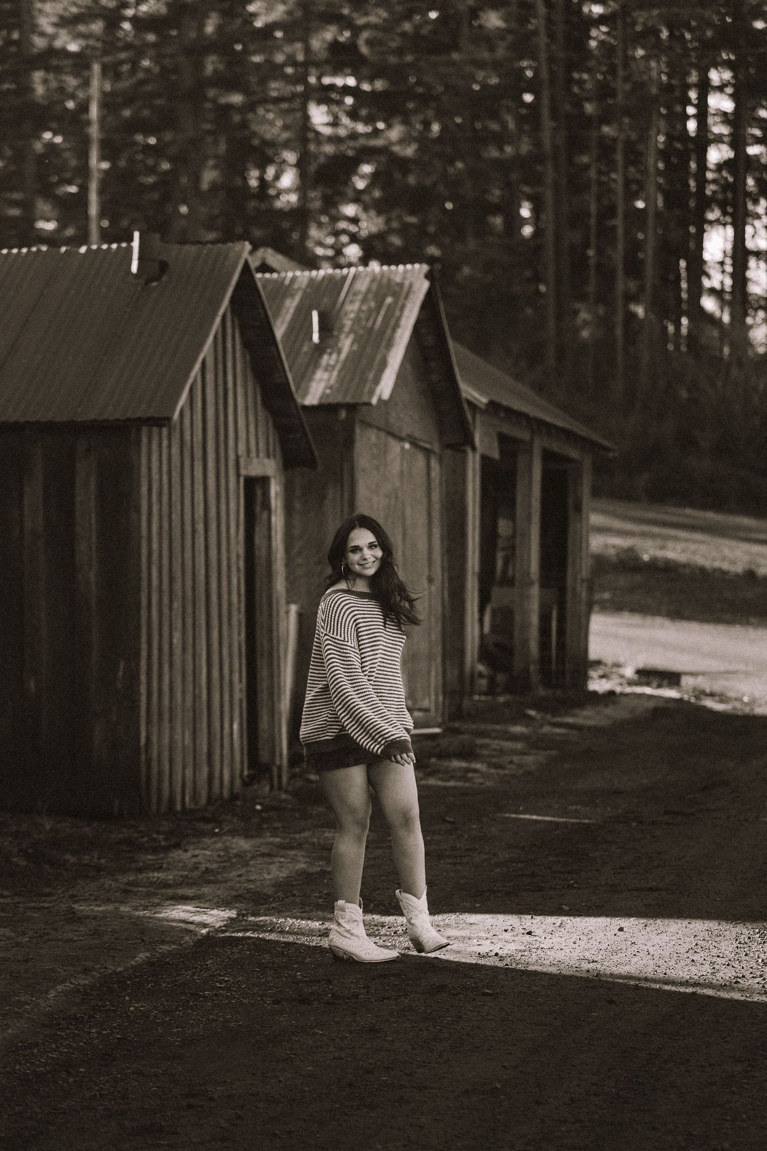 Black and white photo of someone in casual attire walking near wooden cabins in a forest setting.