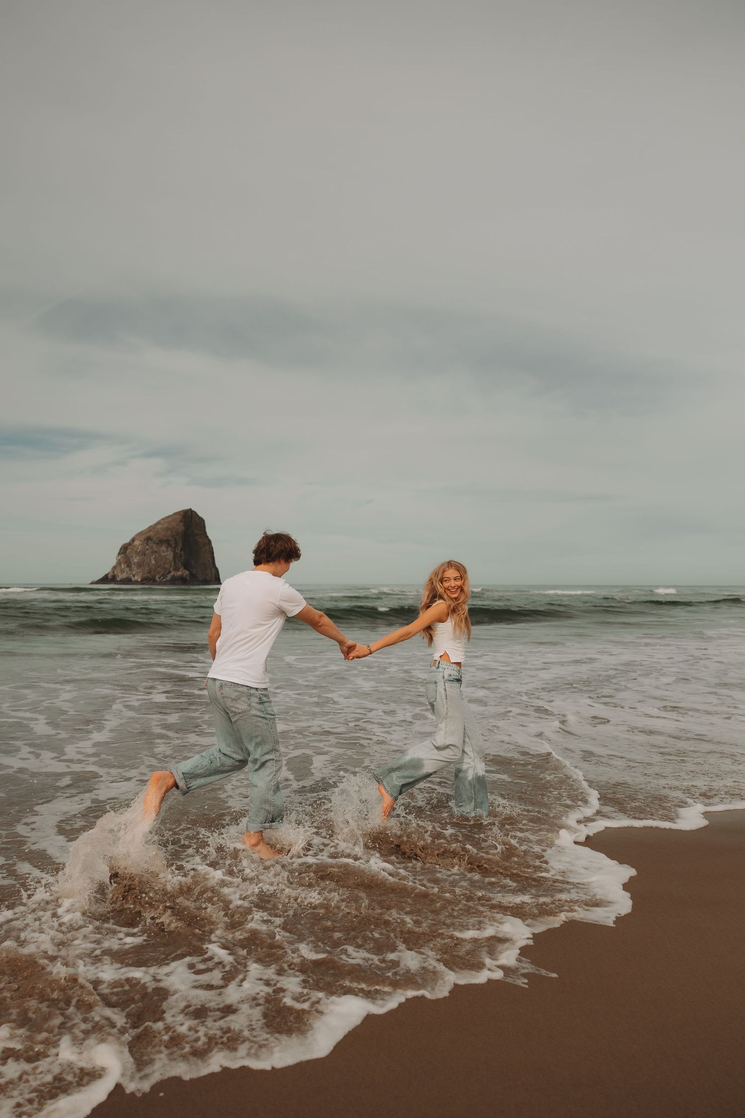 Couple splashes playfully through ocean waves on beach with large sea stack visible.