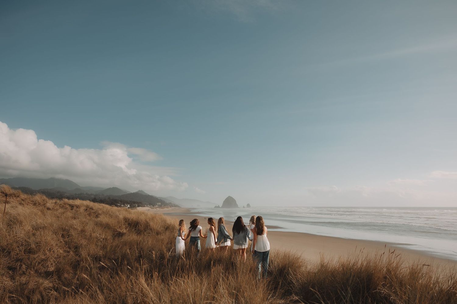 Group of women holding hands walk through coastal dune grass toward the ocean shoreline.