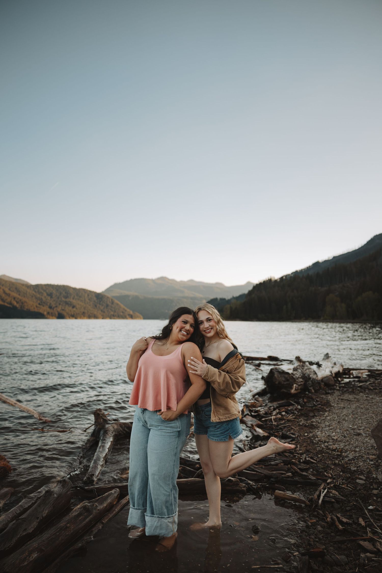 Two young women hugging and smiling on a log-strewn lakeside with mountains at golden hour.