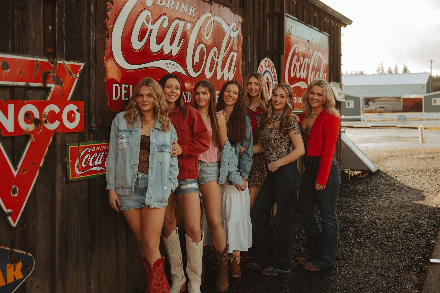Group of women posing by vintage Coca-Cola signs on rustic wooden wall in country western outfits.