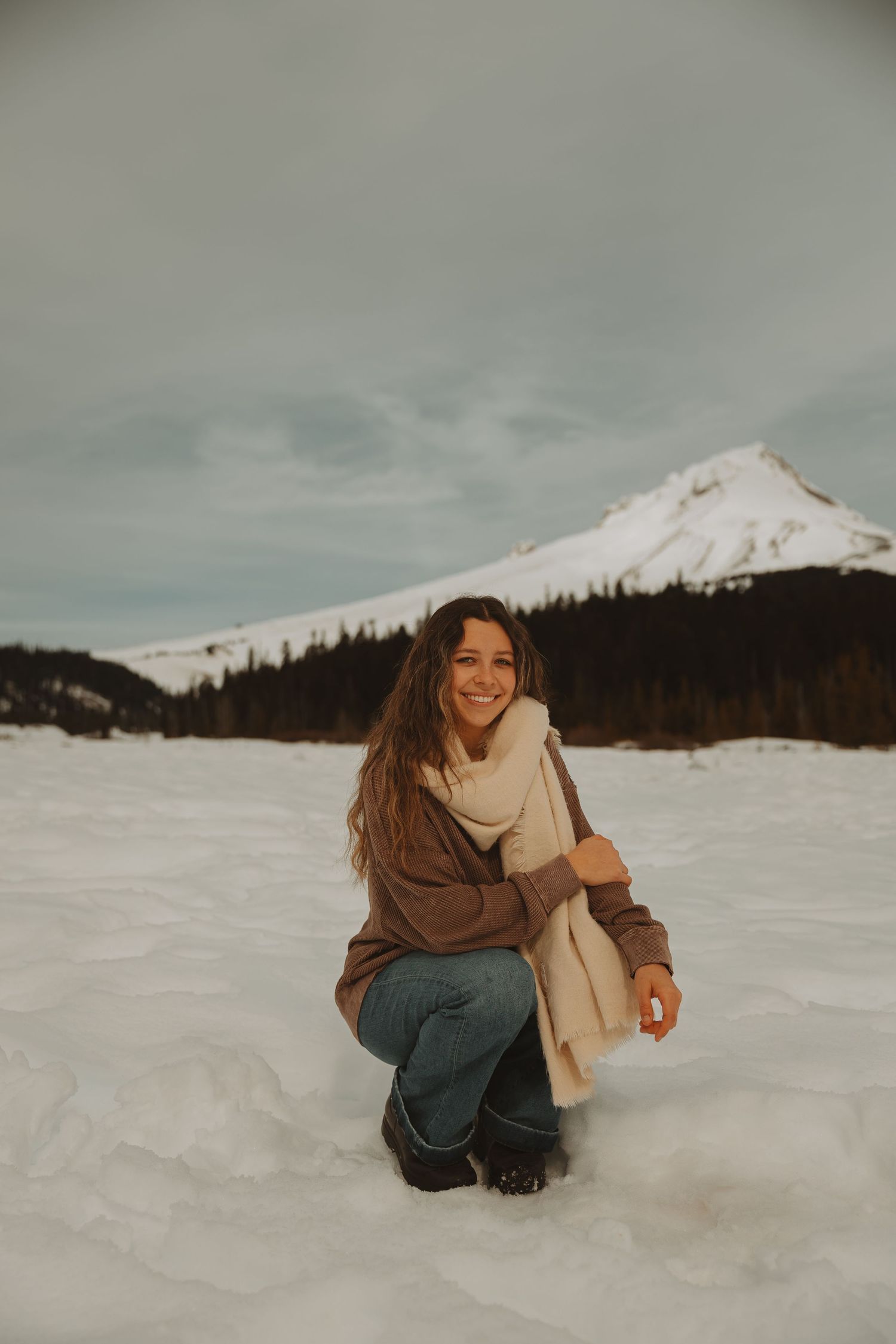 Happy woman in brown hoodie and cream scarf crouches smiling in snowy mountain landscape.