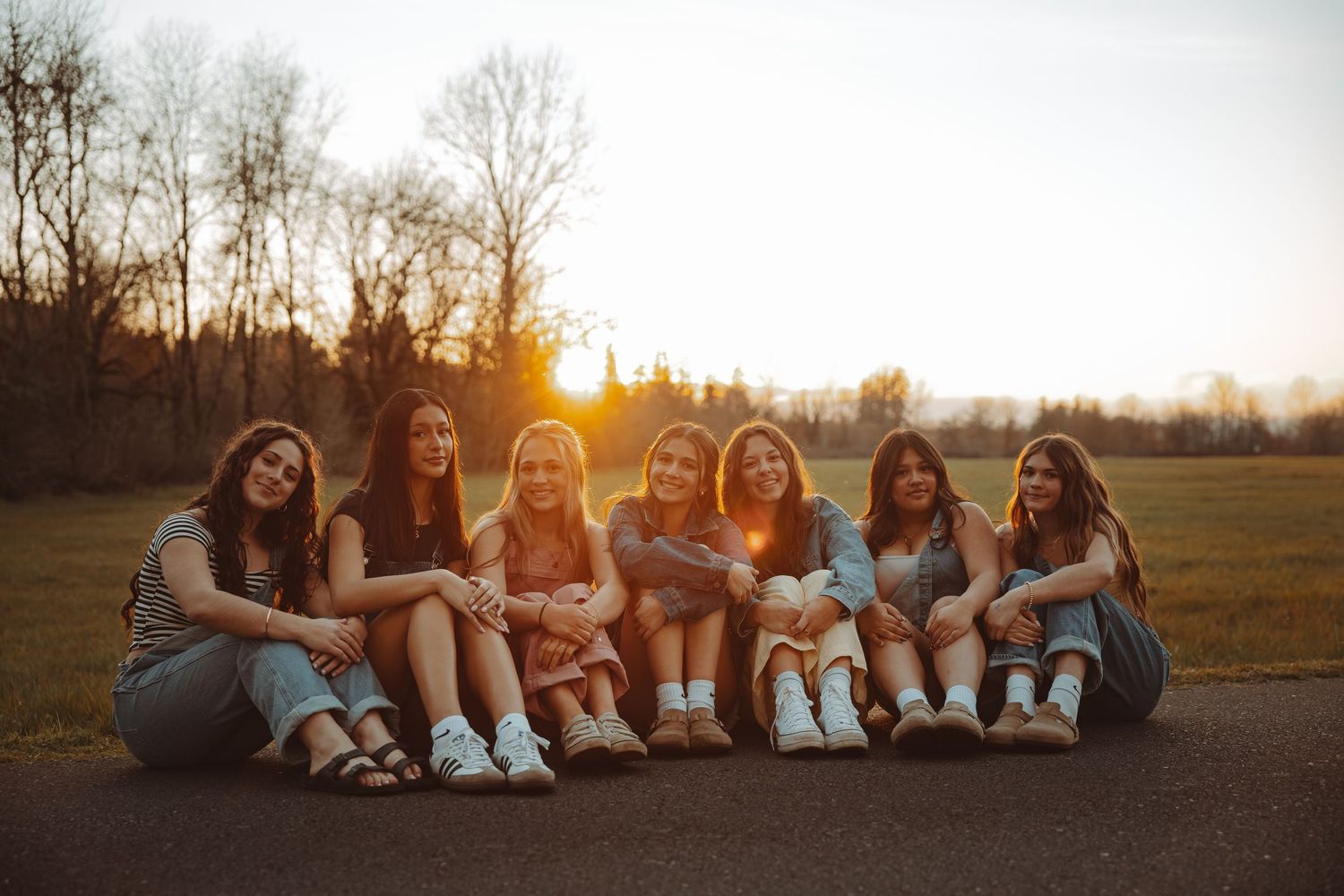 Seven young women sitting together on pavement at sunset, smiling and laughing in a park setting.