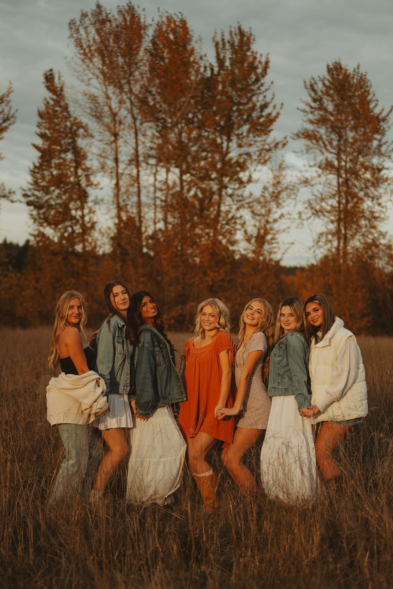 Group of friends in coordinated autumn outfits pose together in a rustic field at sunset.