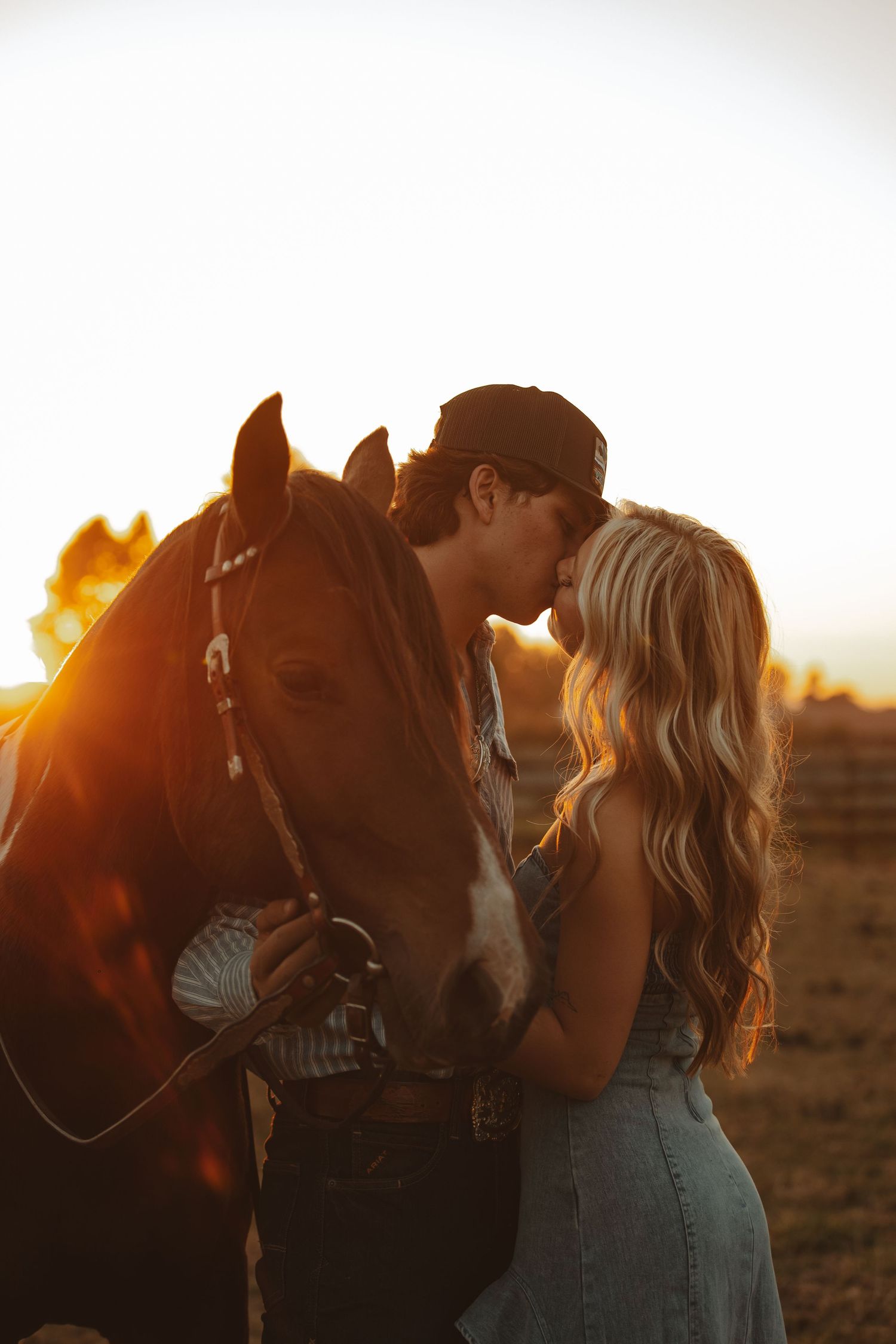 A couple shares a kiss next to their horse during a golden sunset photoshoot.