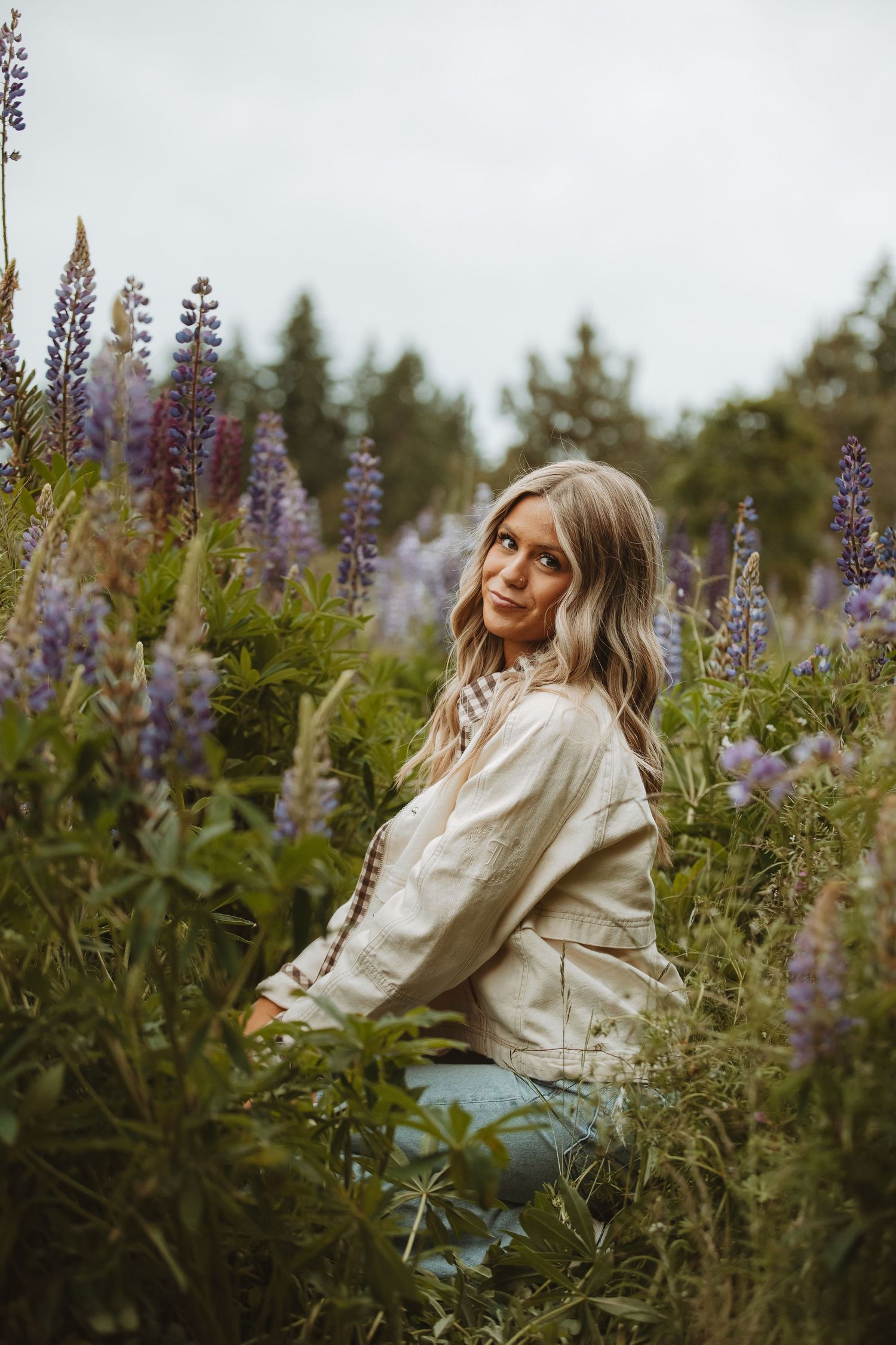Person in cream jacket poses among tall purple lupine flowers in a meadow.