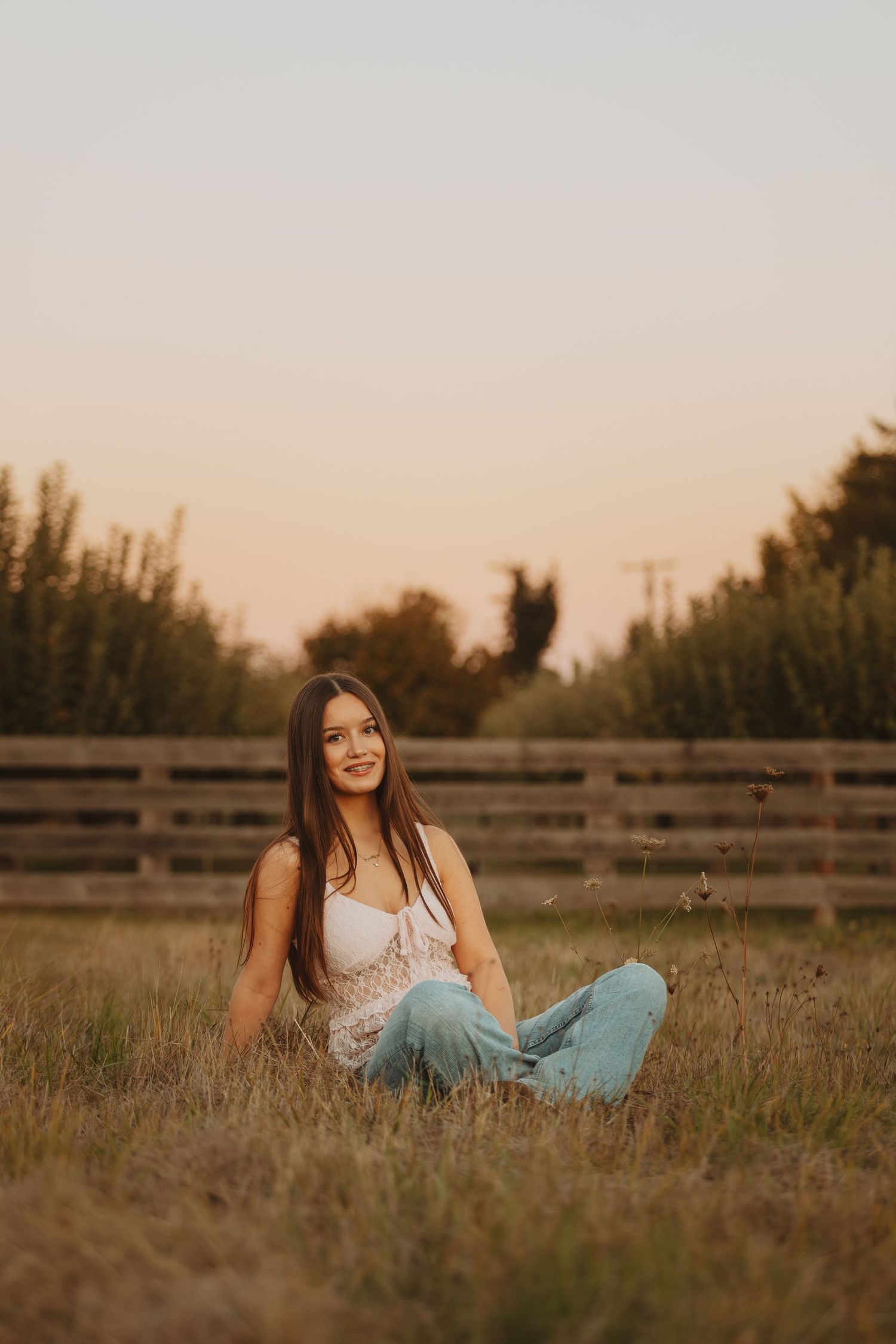 Young woman with long brown hair sits cross-legged in golden field wearing white top and jeans at sunset.