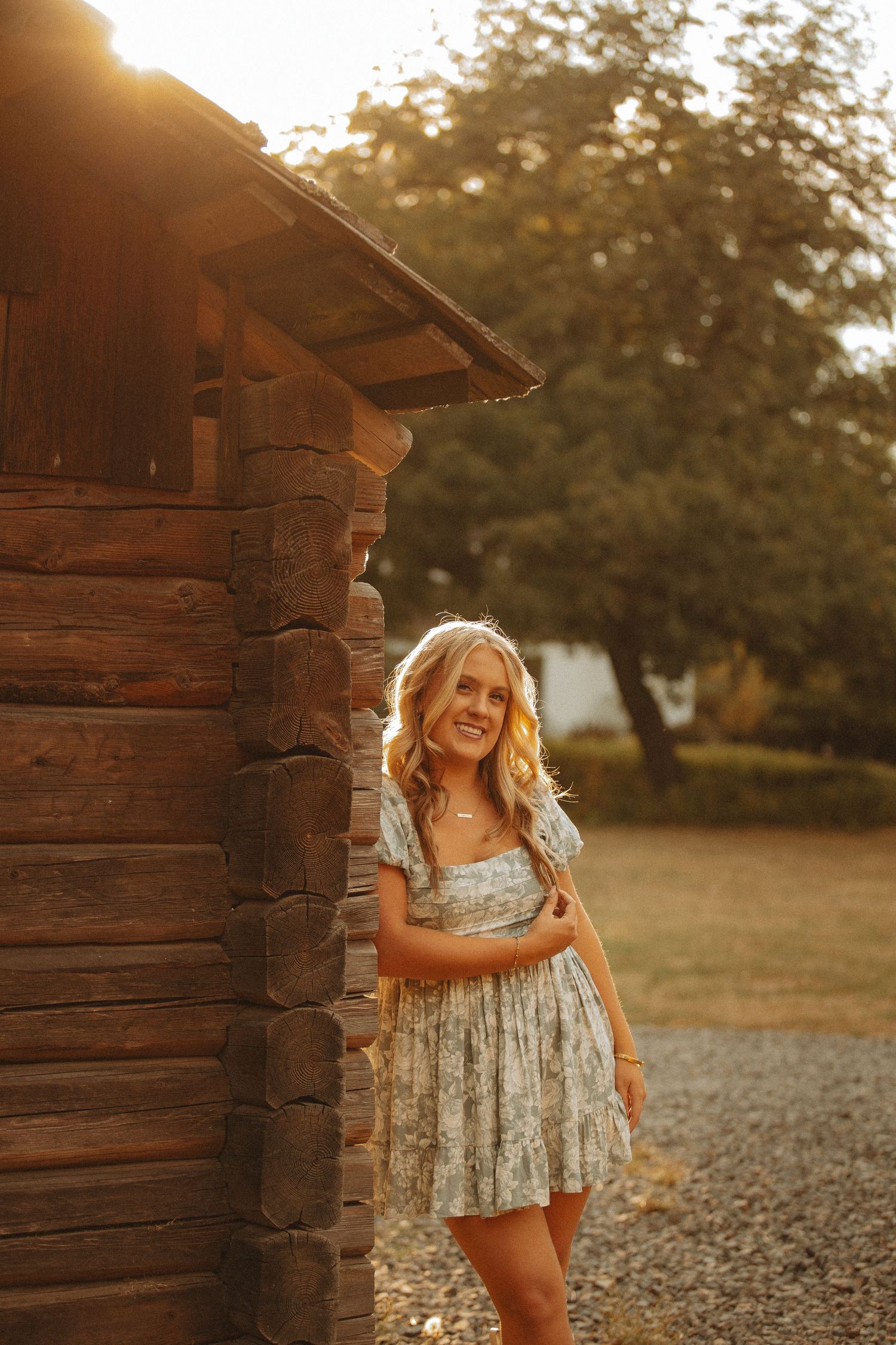 Blonde woman in floral mini dress leaning against rustic log cabin wall during golden sunset.