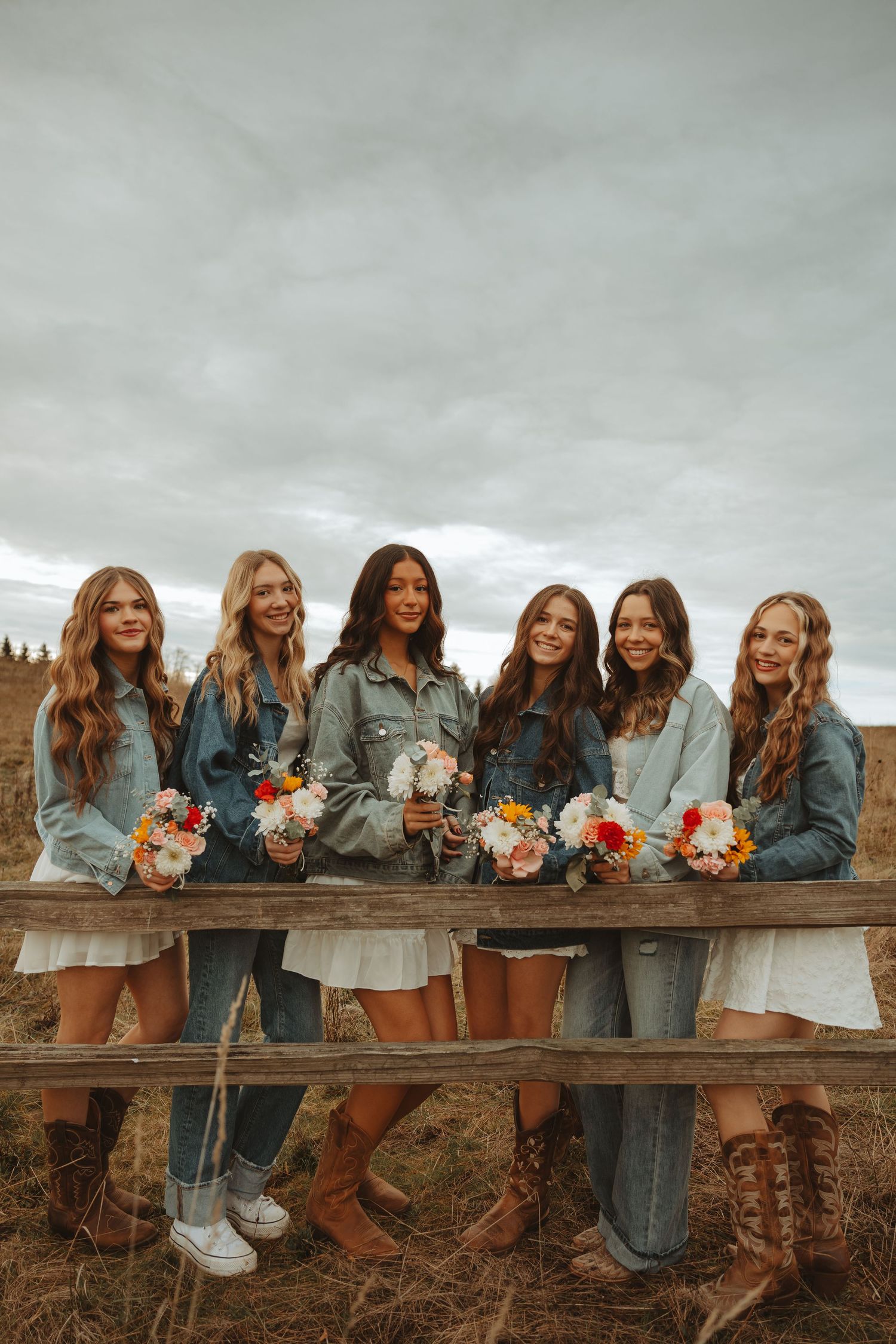 Friends in denim jackets lean on a wooden fence holding colorful bouquets against a cloudy sky.