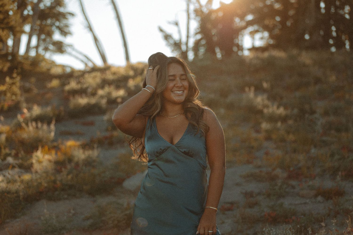 Woman in teal dress laughing outdoors at golden hour, hand on head, surrounded by desert shrubs and warm sunlight.