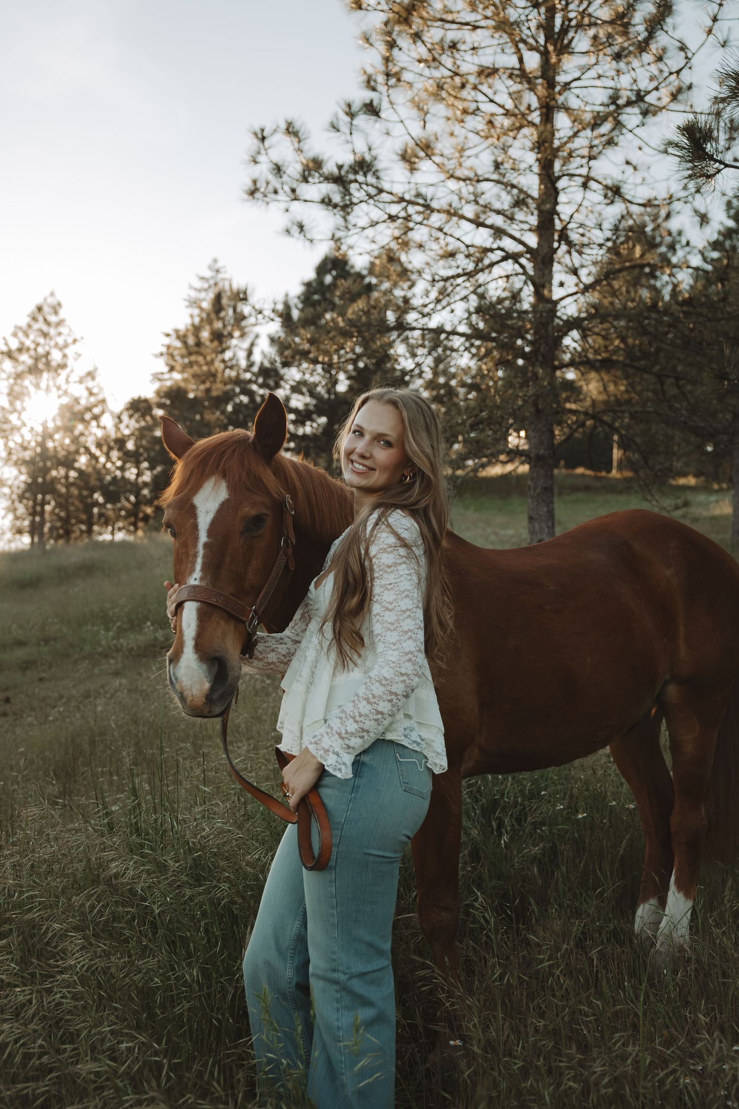 Young blonde woman in white lace top and jeans smiling beside a chestnut horse in a sunlit green field.