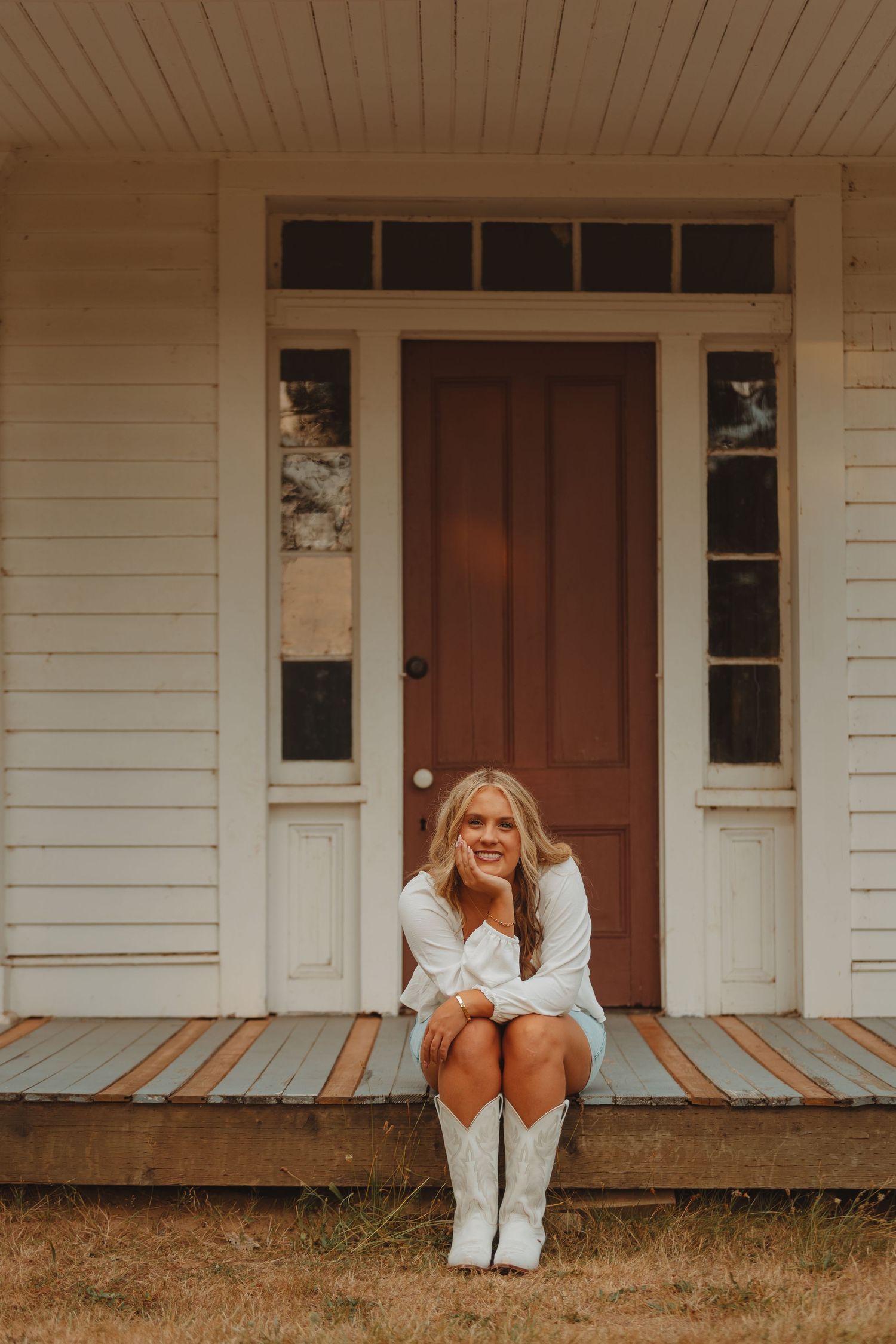 Smiling blonde woman in white dress and cowboy boots sitting on rustic porch with red door.