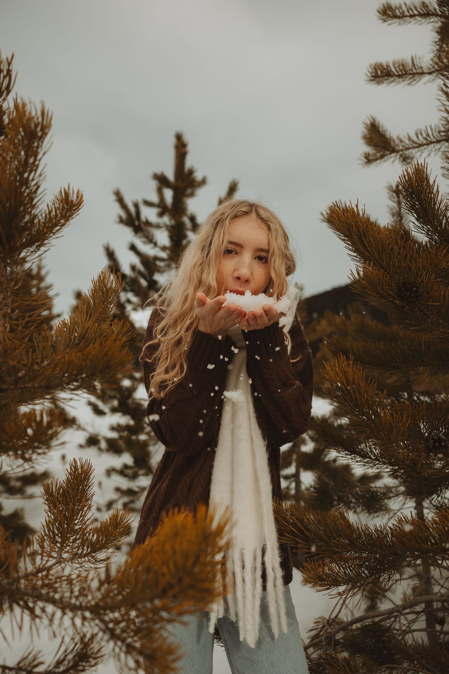 Blonde woman blowing snow from hands surrounded by pine trees in a winter forest setting.