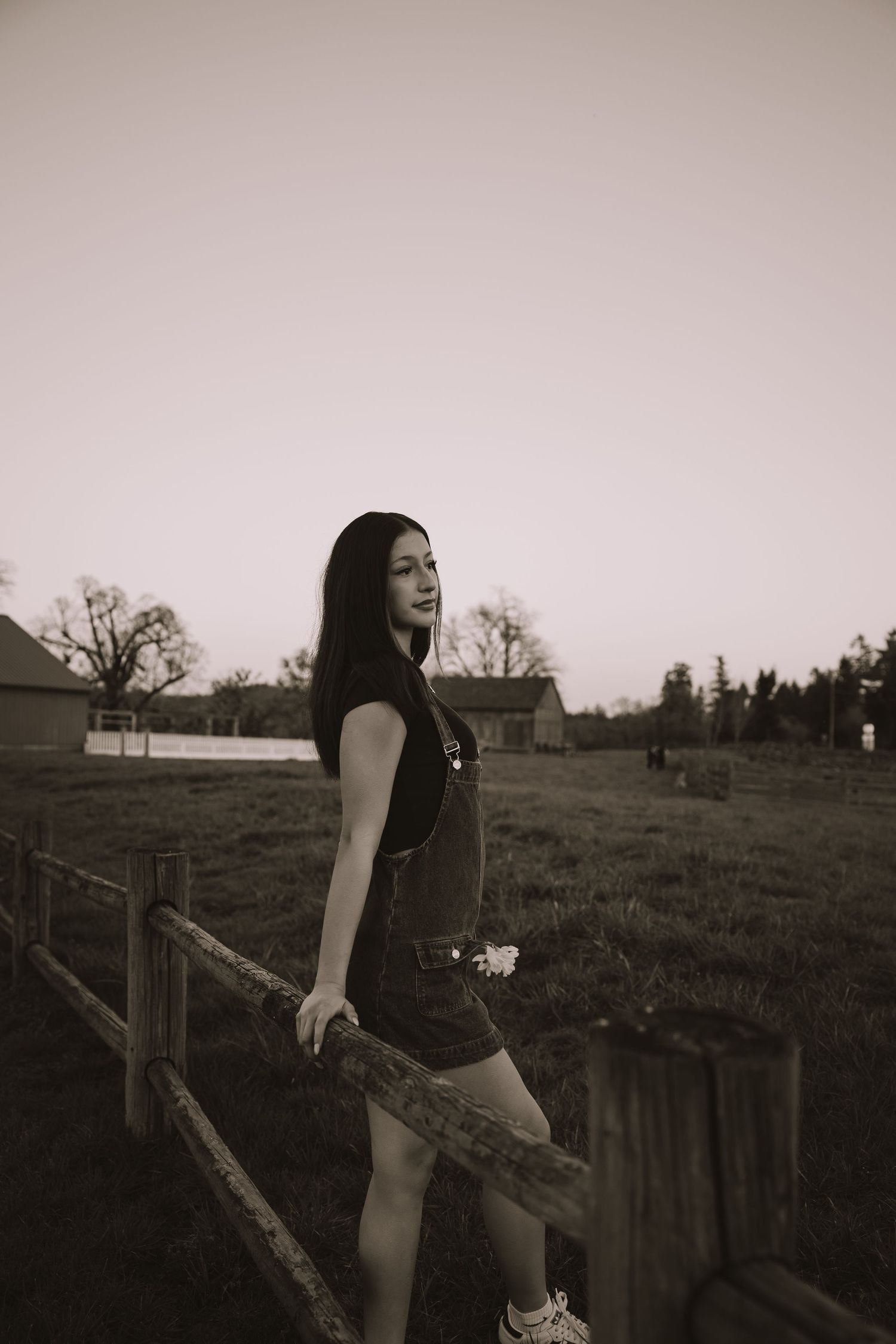 Young woman in overalls posing by a wooden fence in a rural field at dusk in black and white.