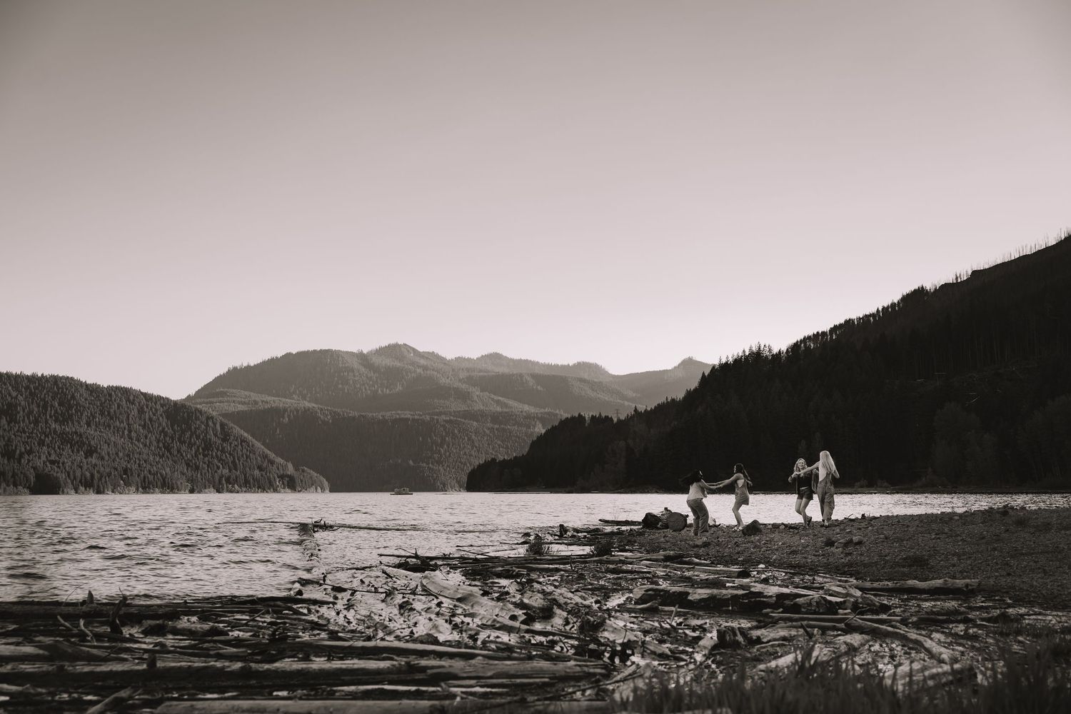 Black and white photo of people walking along a rocky shoreline with mountain lake backdrop.