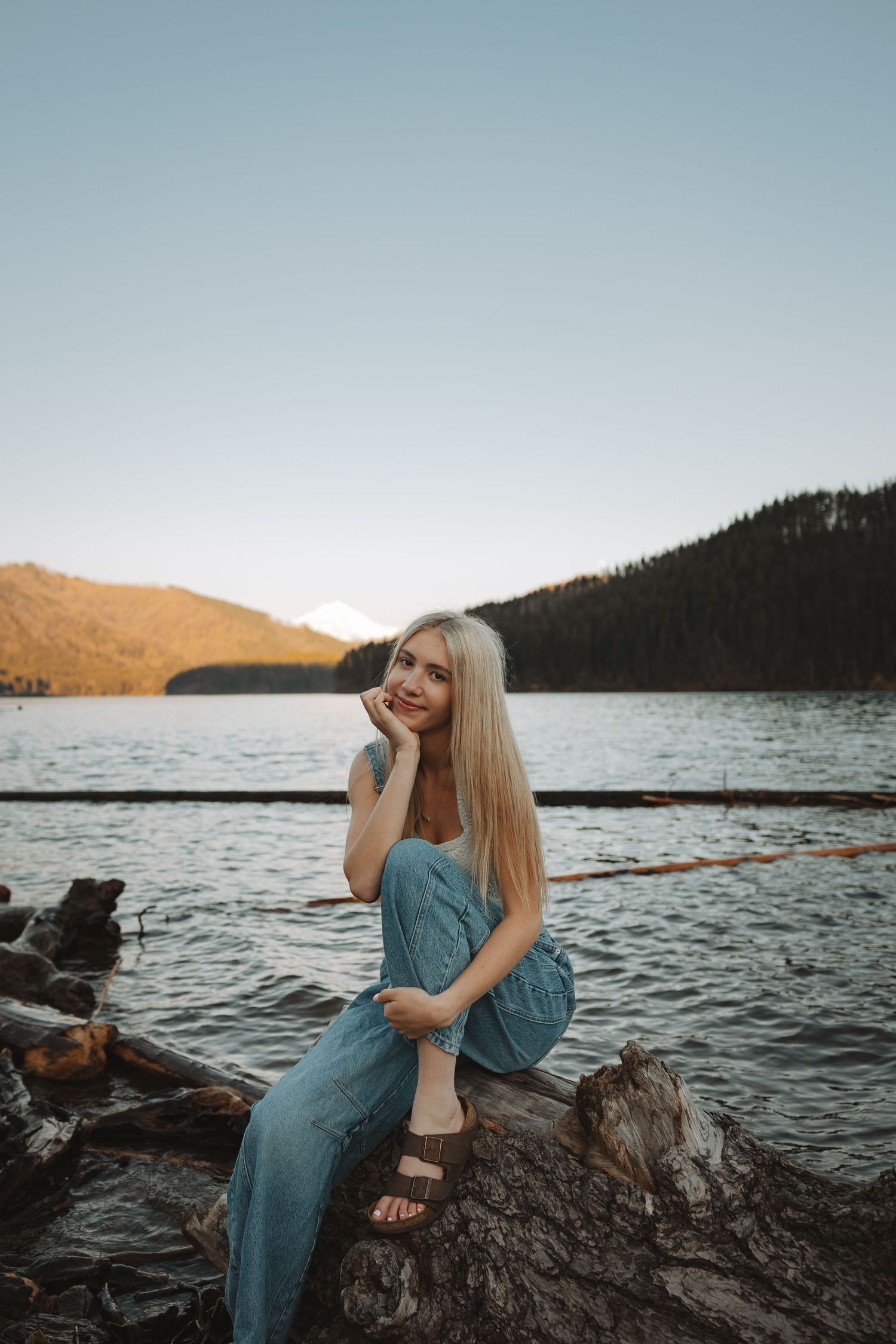 Blonde woman in blue dress sitting on driftwood logs by a serene mountain lake at sunset.