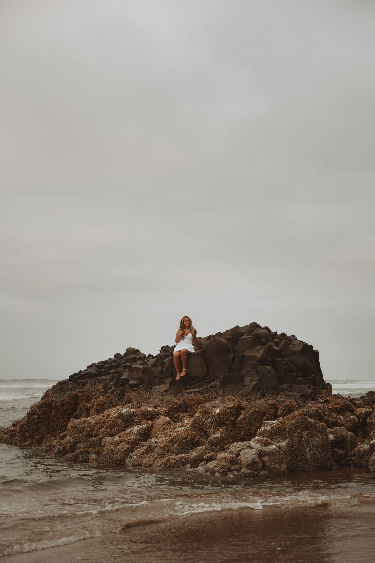 Woman in white dress sitting atop rocky formation on overcast beach with ocean waves below.