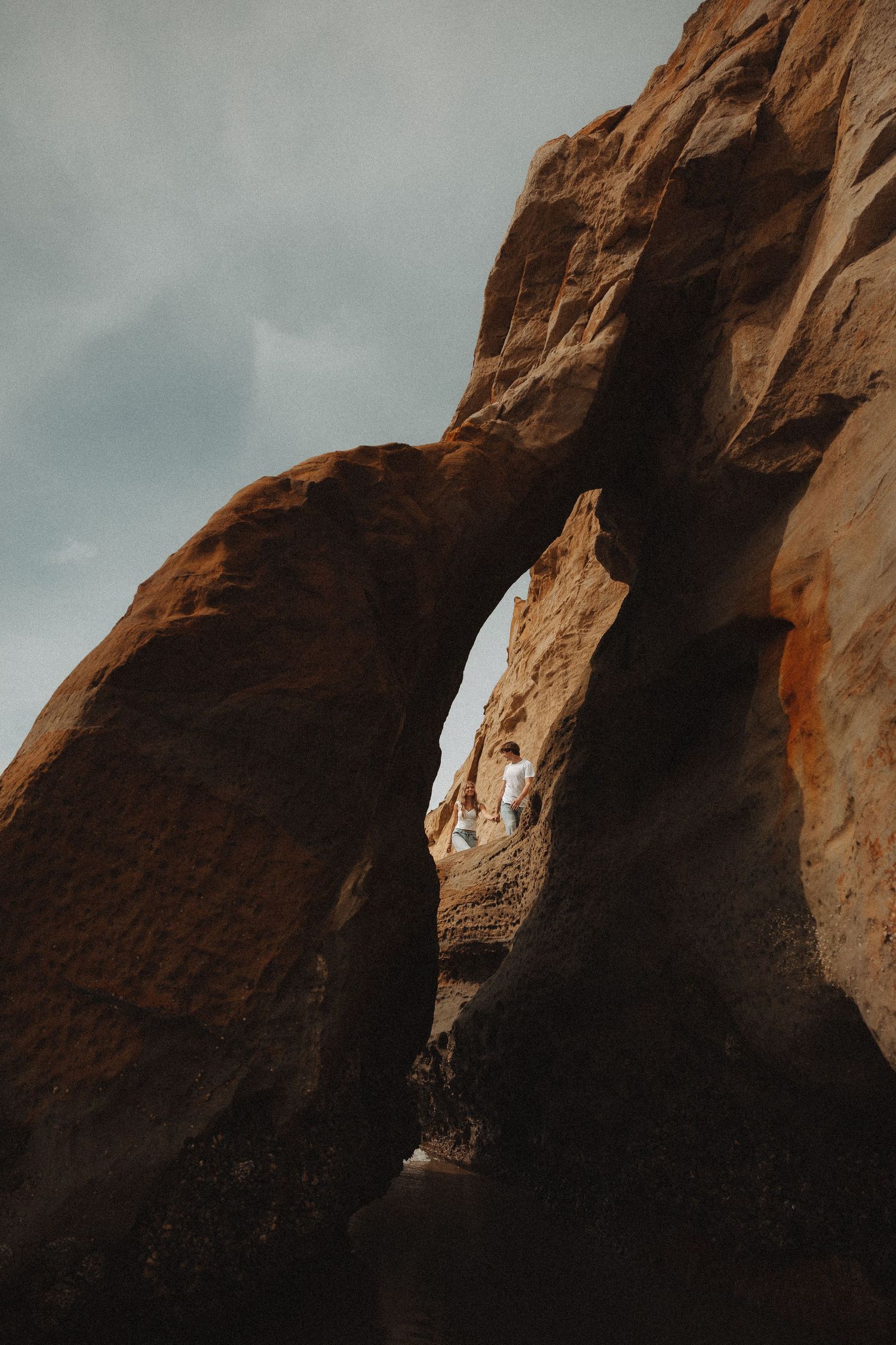 A couple stands beneath a dramatic natural sandstone arch in a desert landscape under a moody overcast sky.