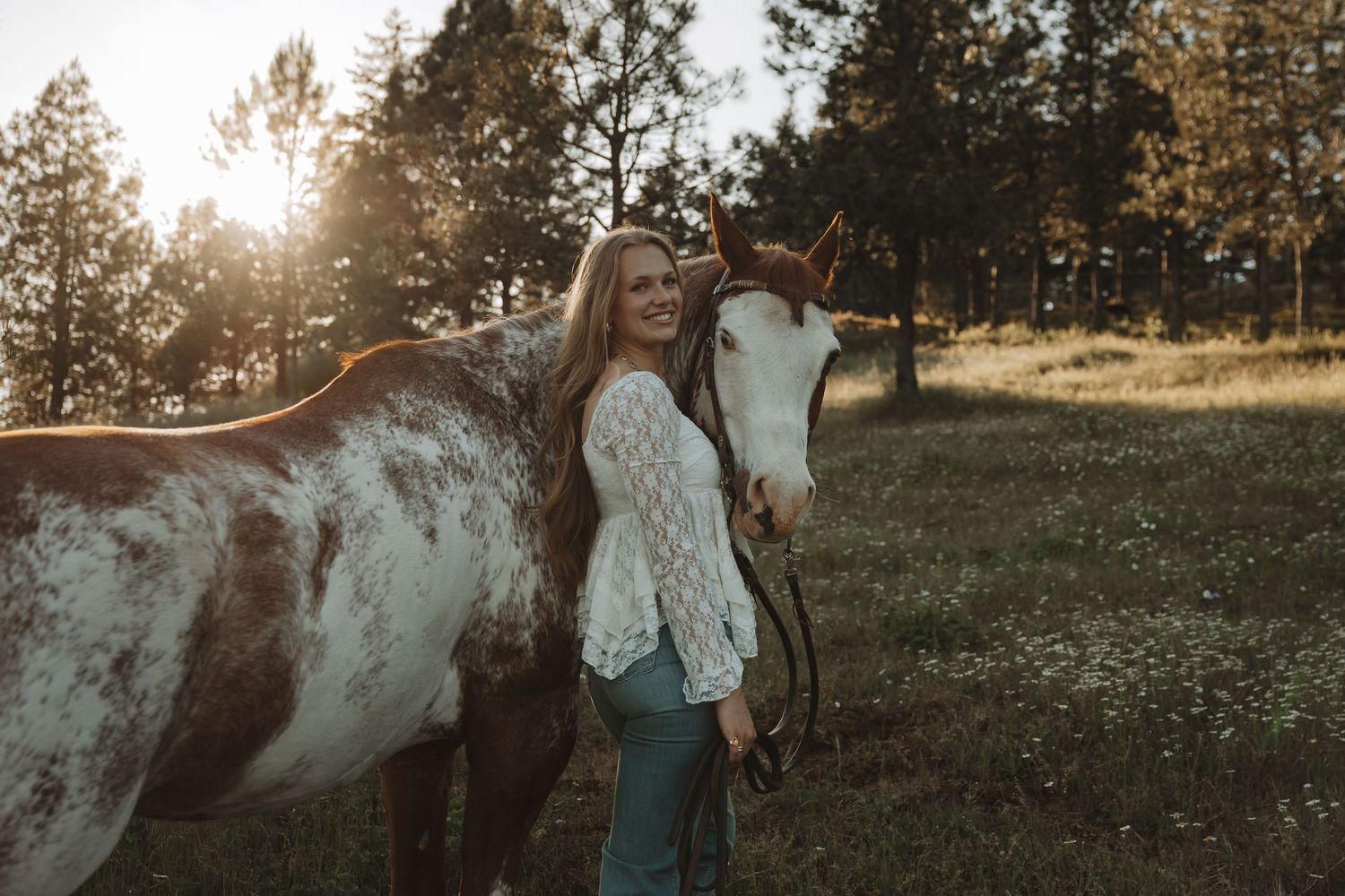 A smiling blonde woman in a white lace top poses with a brown and white horse in a sunlit meadow.