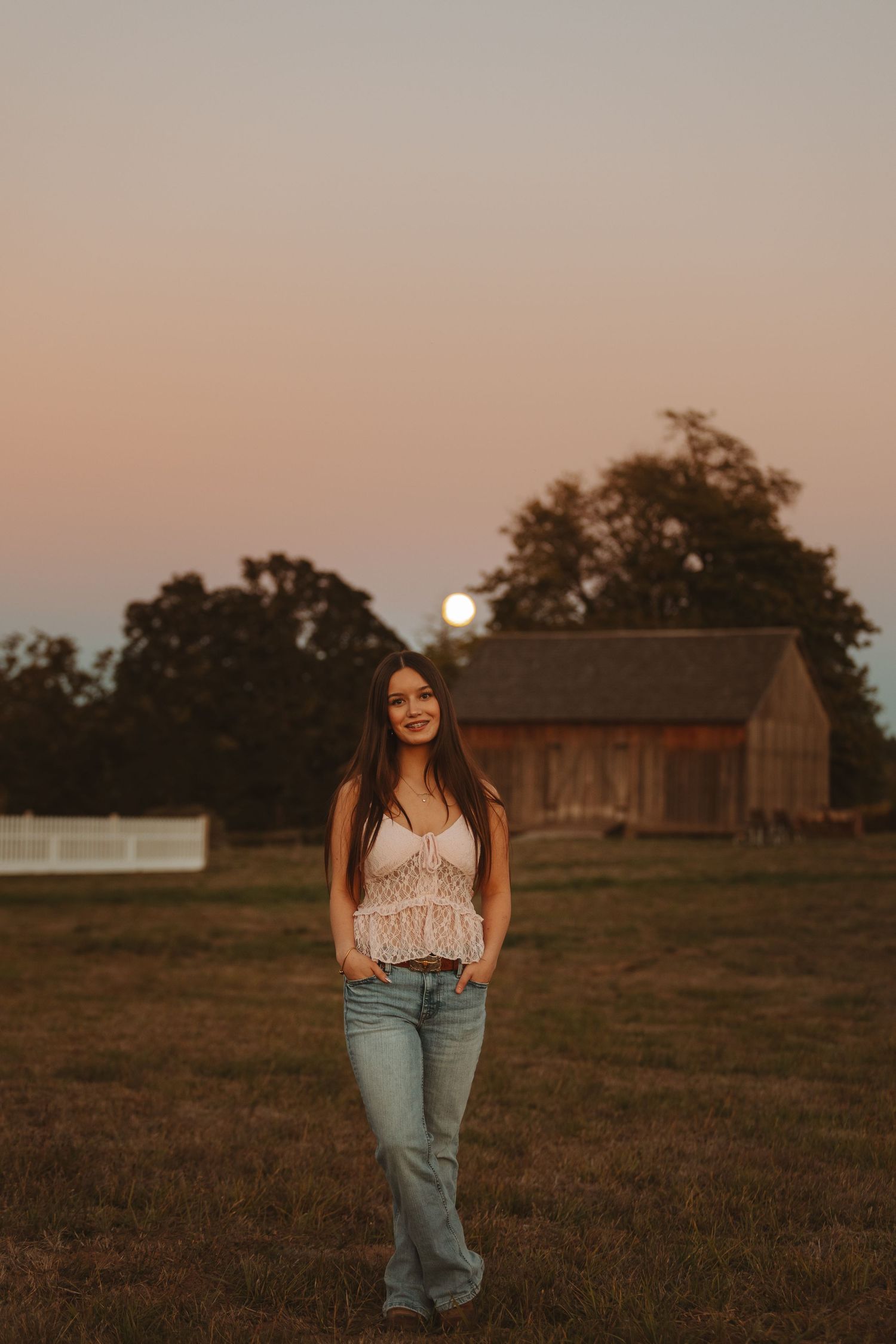 Young woman standing in a rural field at dusk, wearing a white lace top and flared jeans with a barn and full moon behind her.
