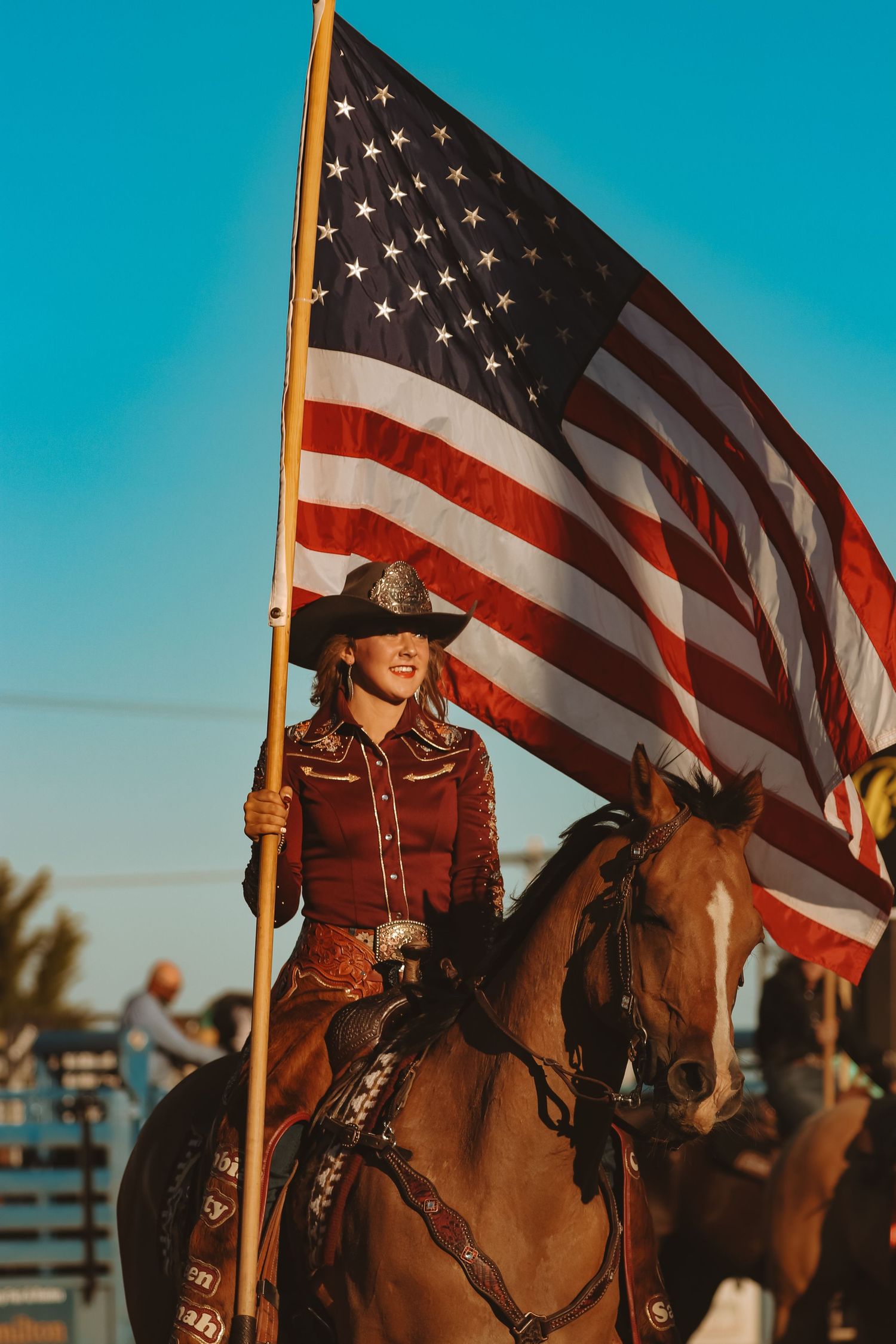 A rider carries the American flag while mounted on a brown horse during a rodeo ceremony.