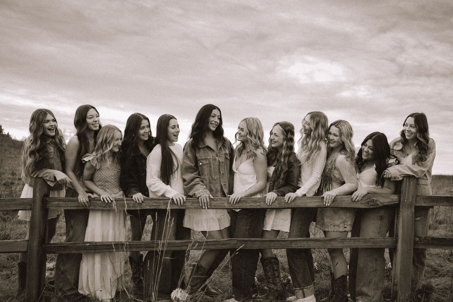 A group of people in bohemian-style dresses and long hair lean against a rustic wooden fence in a black and white photograph.