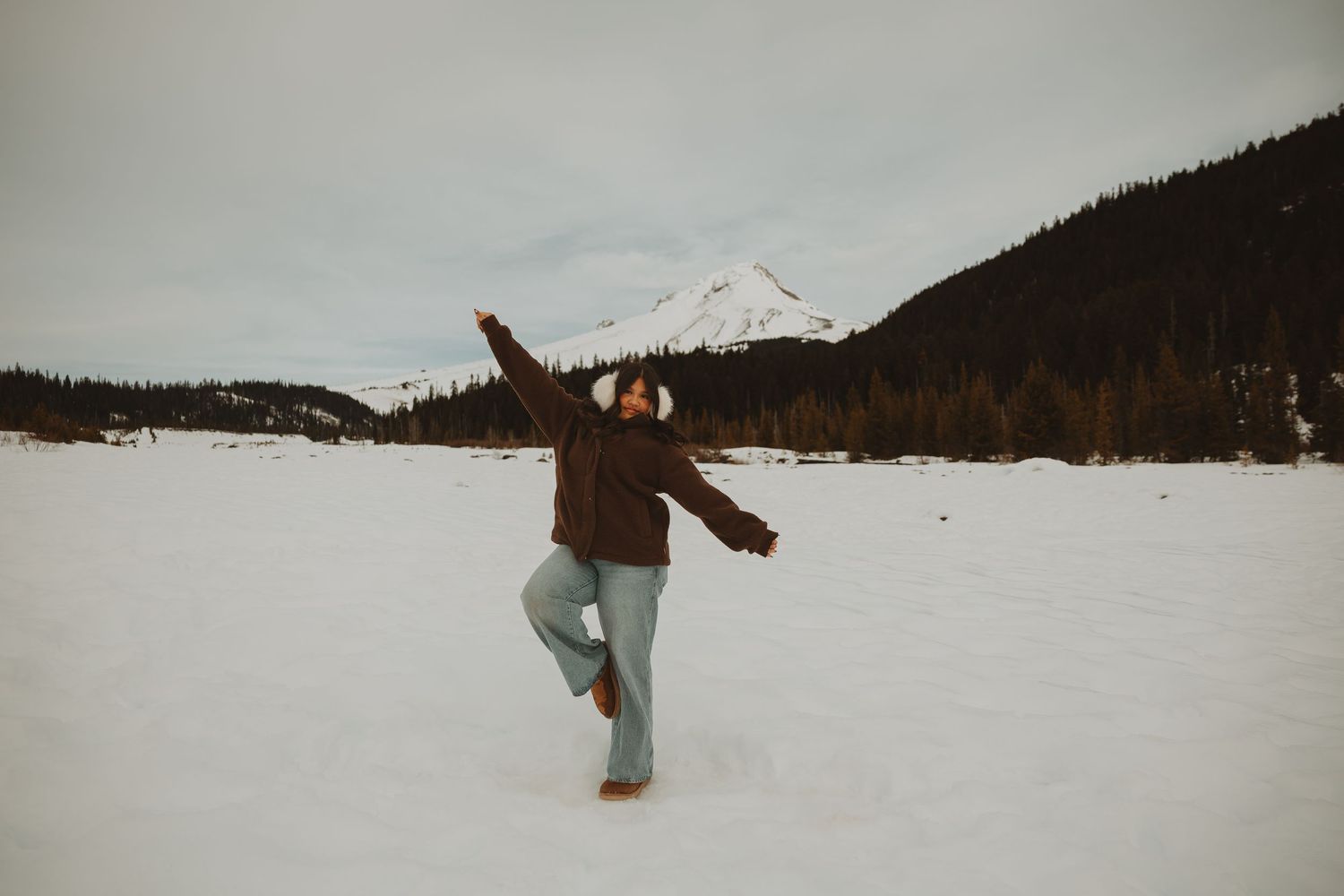 Woman in brown jacket balances on one leg with arms raised on a snowy field near mountains.