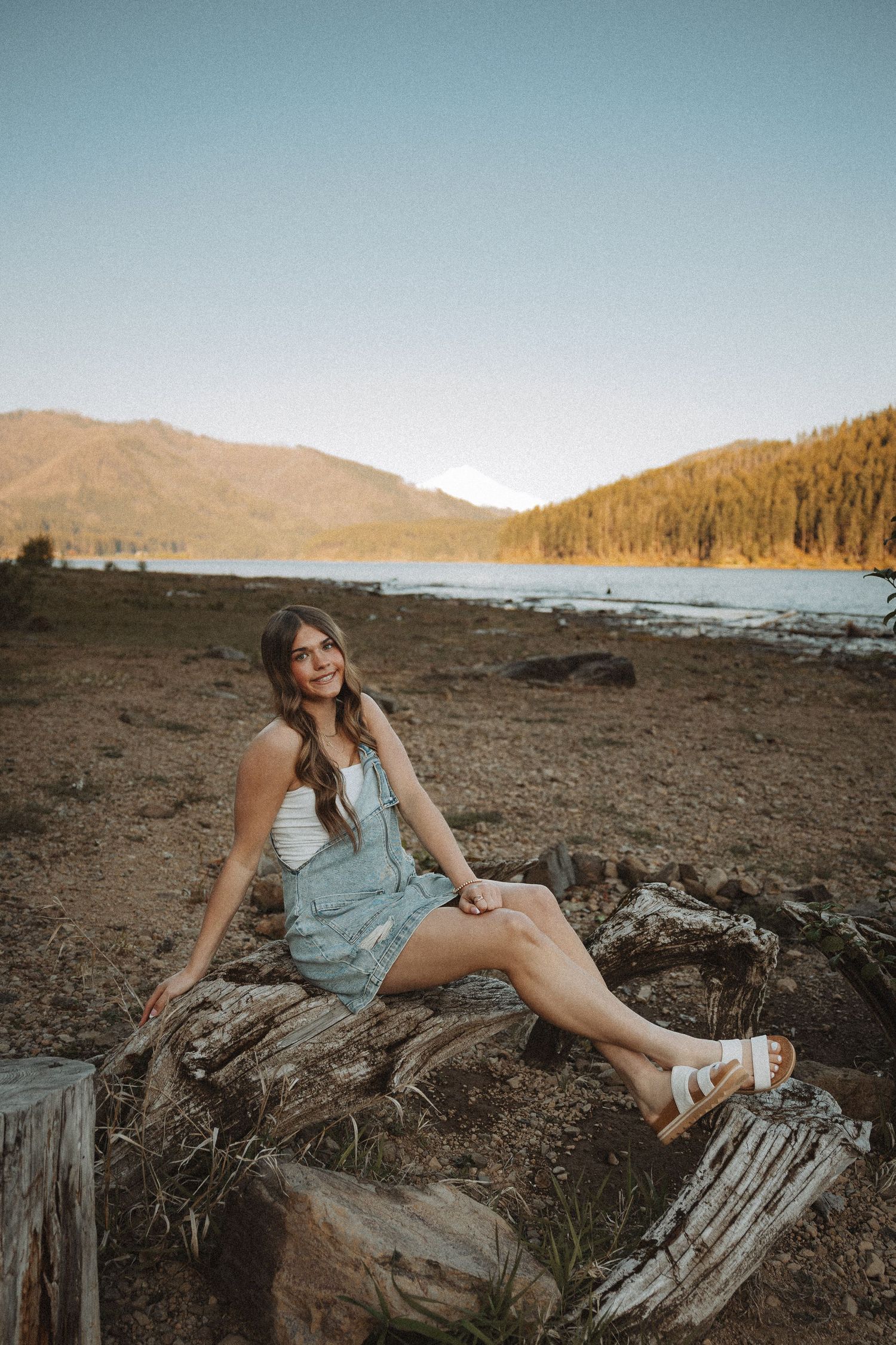 Smiling brunette woman in blue romper and white sandals posing on logs near a scenic mountain lake.
