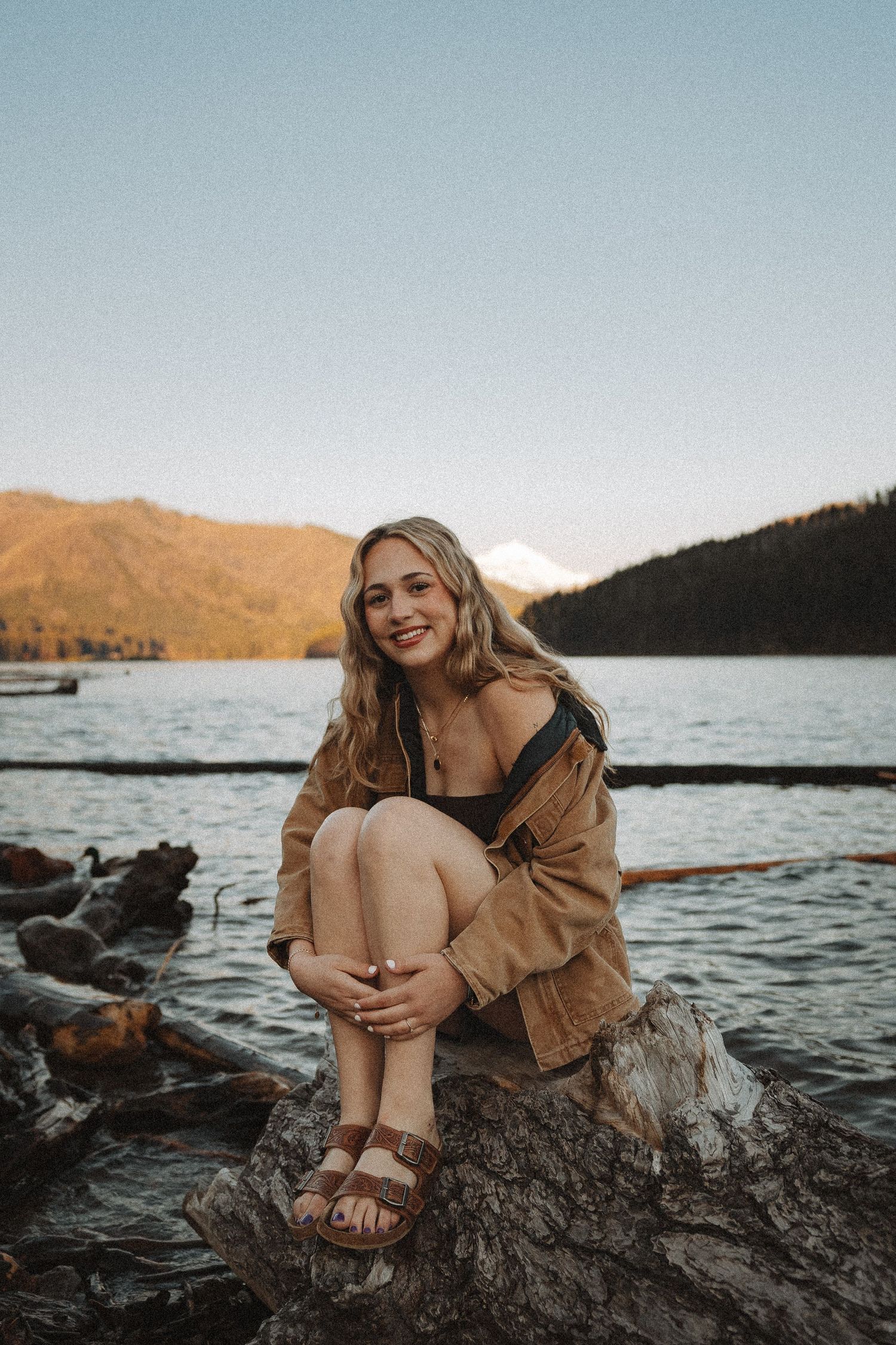 Young woman in brown jacket sitting on driftwood by a mountain lake at sunset.