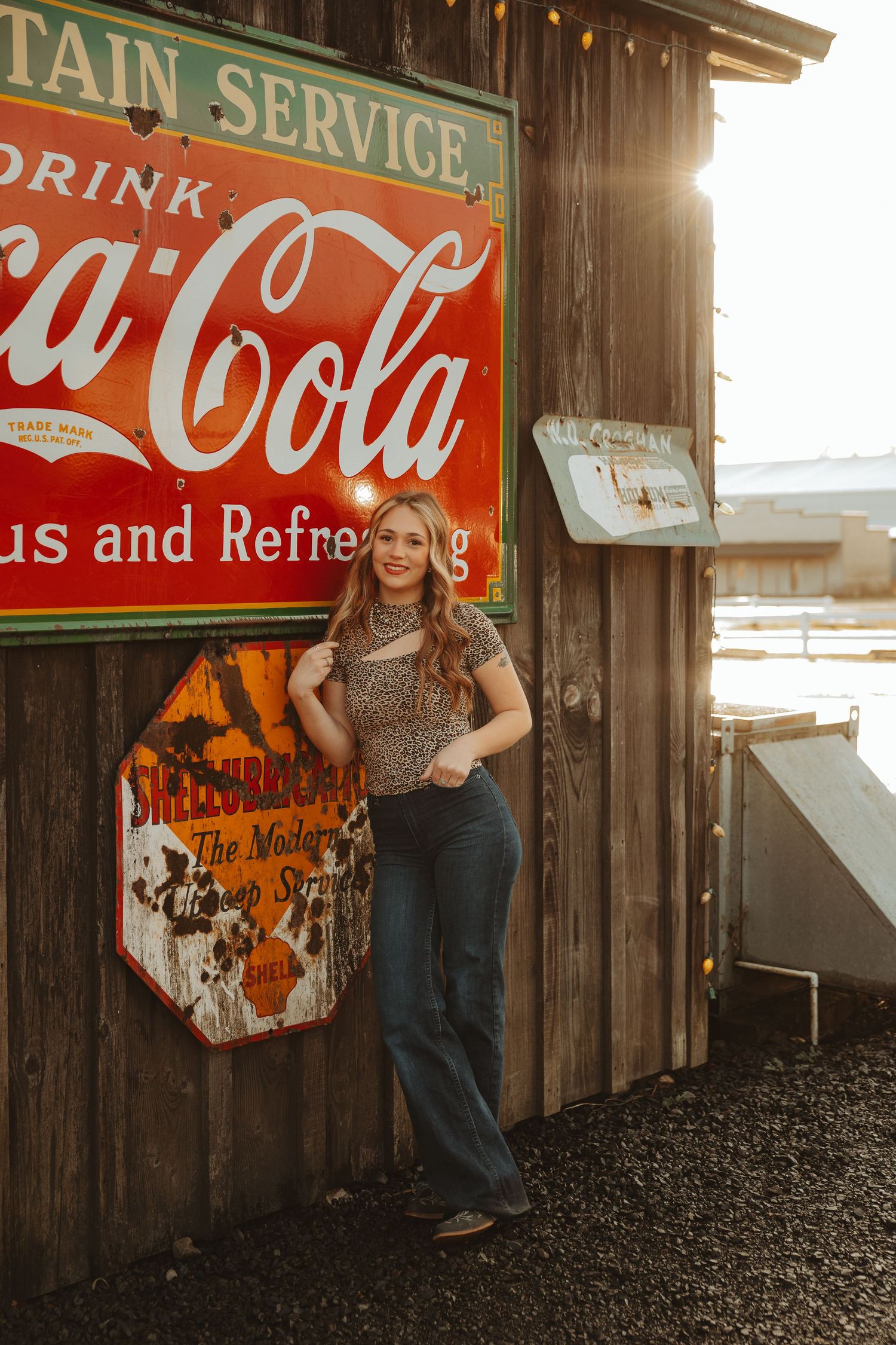 Young woman in leopard print top and flare jeans posing by vintage Coca-Cola sign on rustic wooden building at sunset.