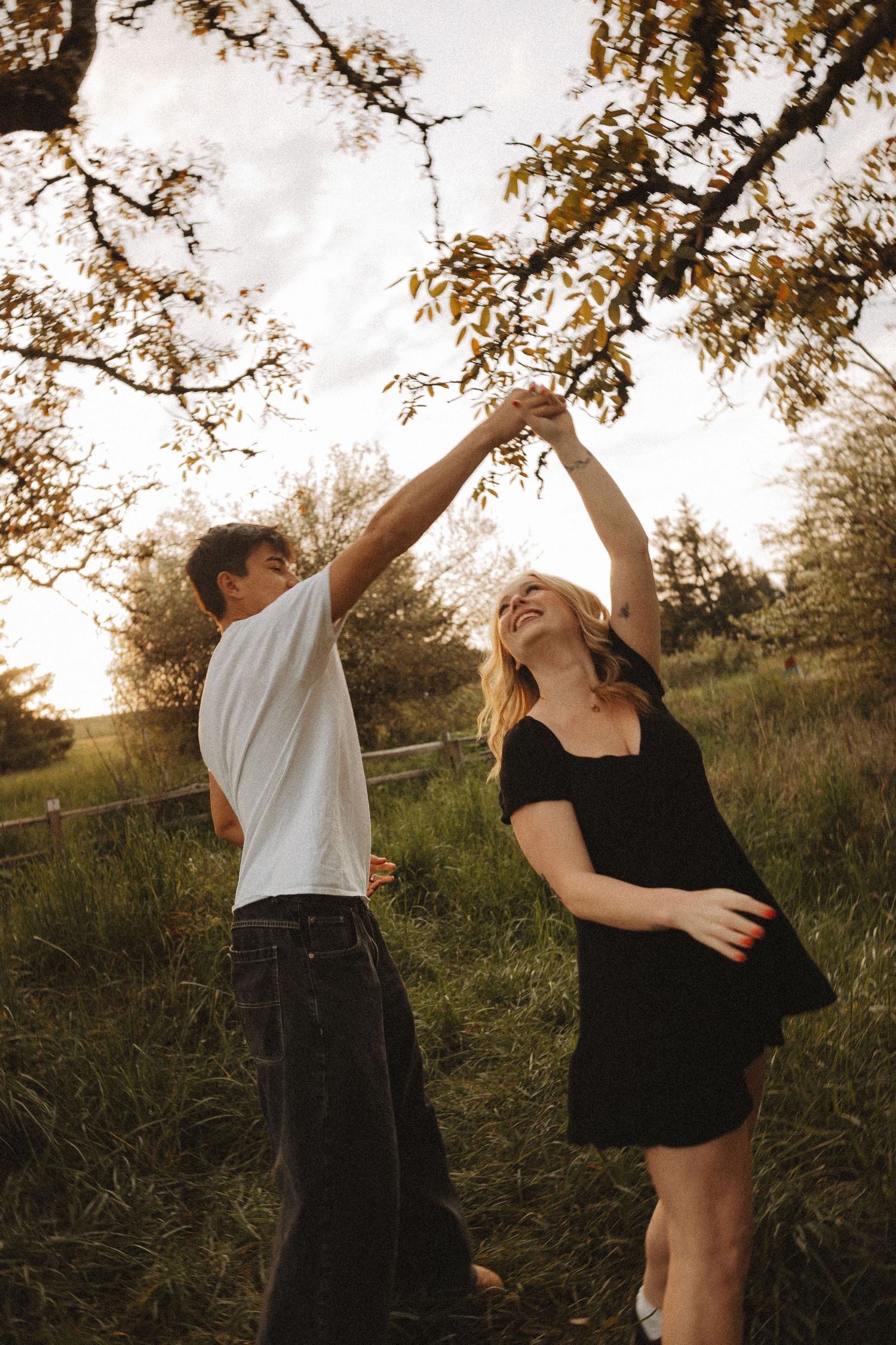 A joyful couple dancing outdoors at sunset, man in white tee spinning woman in black dress under a tree.