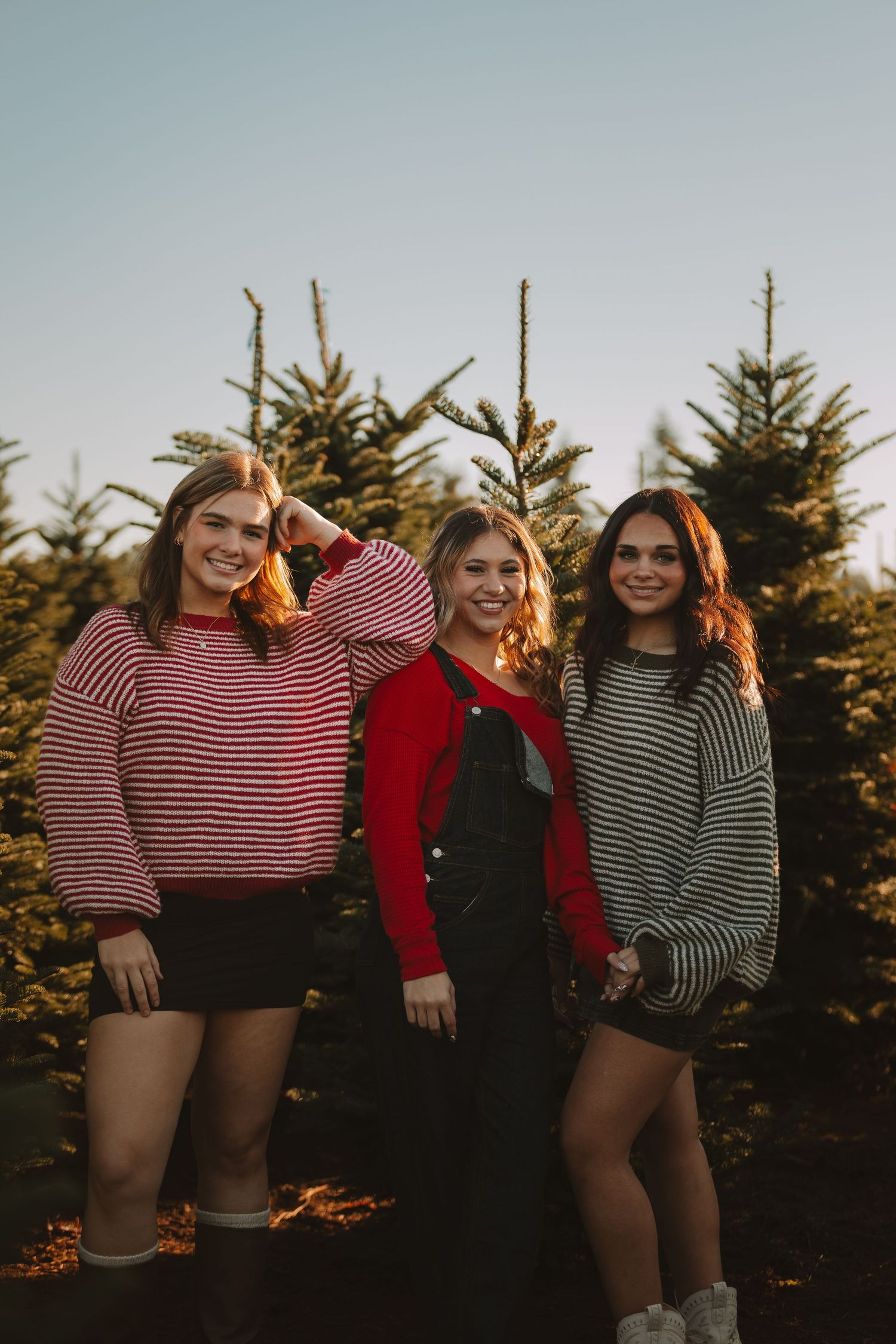 Friends in striped shirts and casual wear pose together in front of evergreen trees during golden hour.