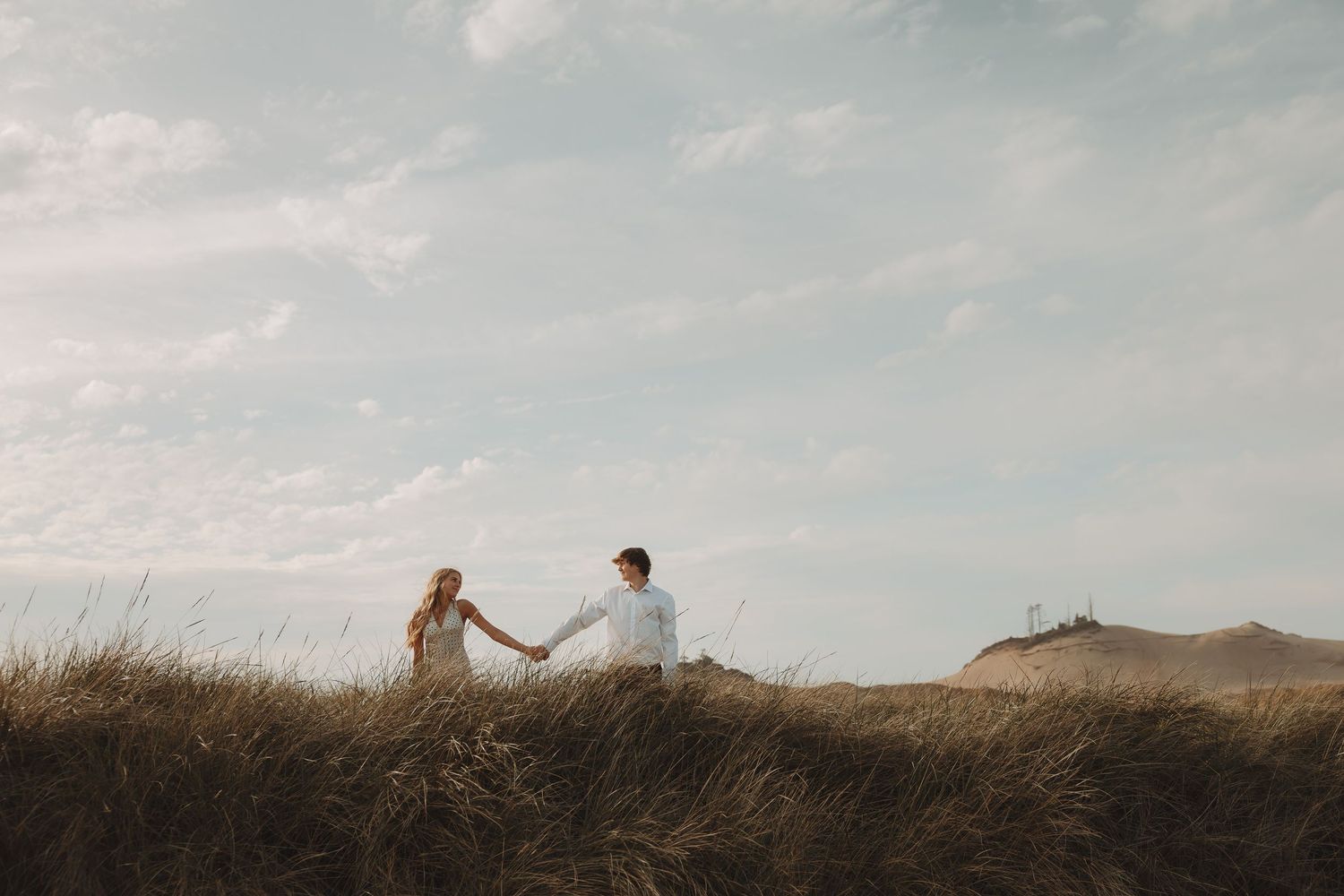 Couple holding hands walking through golden dunes under a cloudy sky in a romantic outdoor engagement photo.