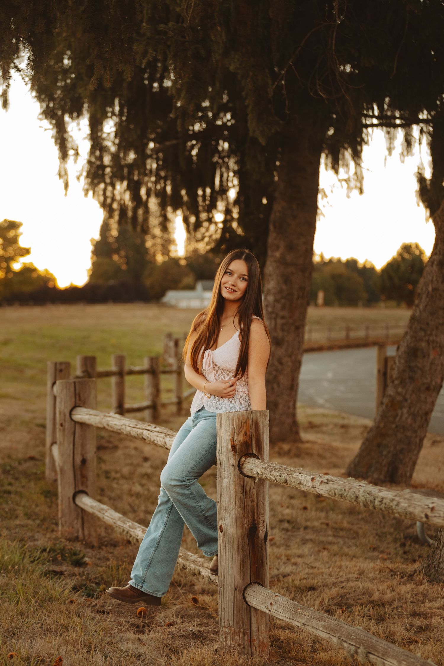 Young woman in jeans and white top leaning on rustic wooden fence at golden hour on a farm.