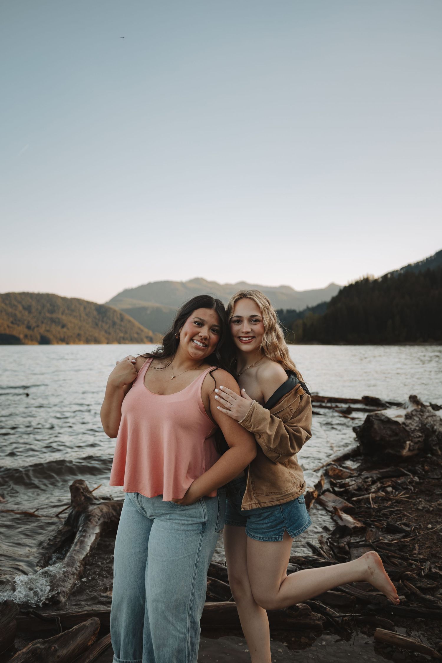 Two young women hugging and smiling on rocky lakeshore with forested mountains in background at sunset.