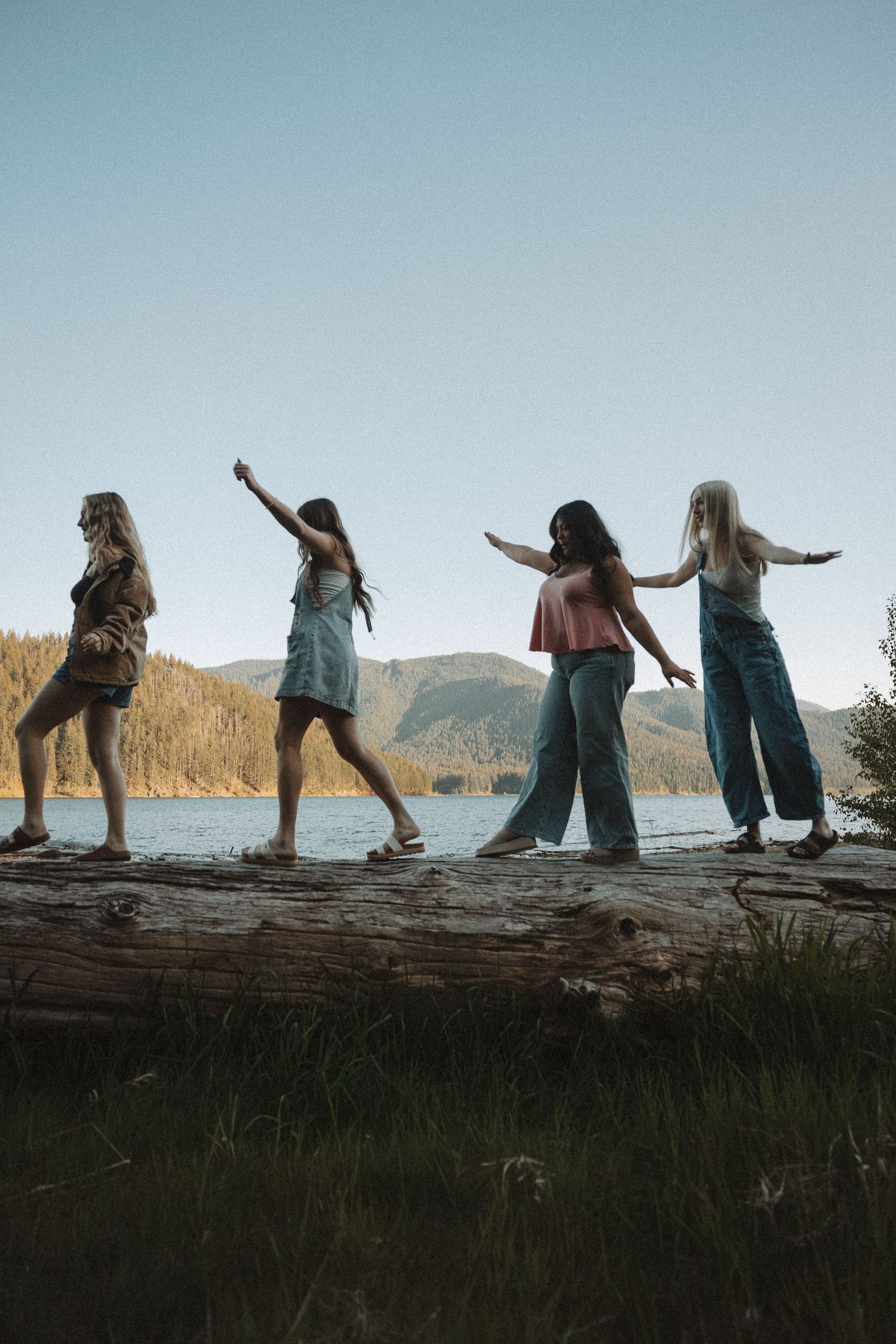 Four women balance on a fallen log near a mountain lake at golden hour.