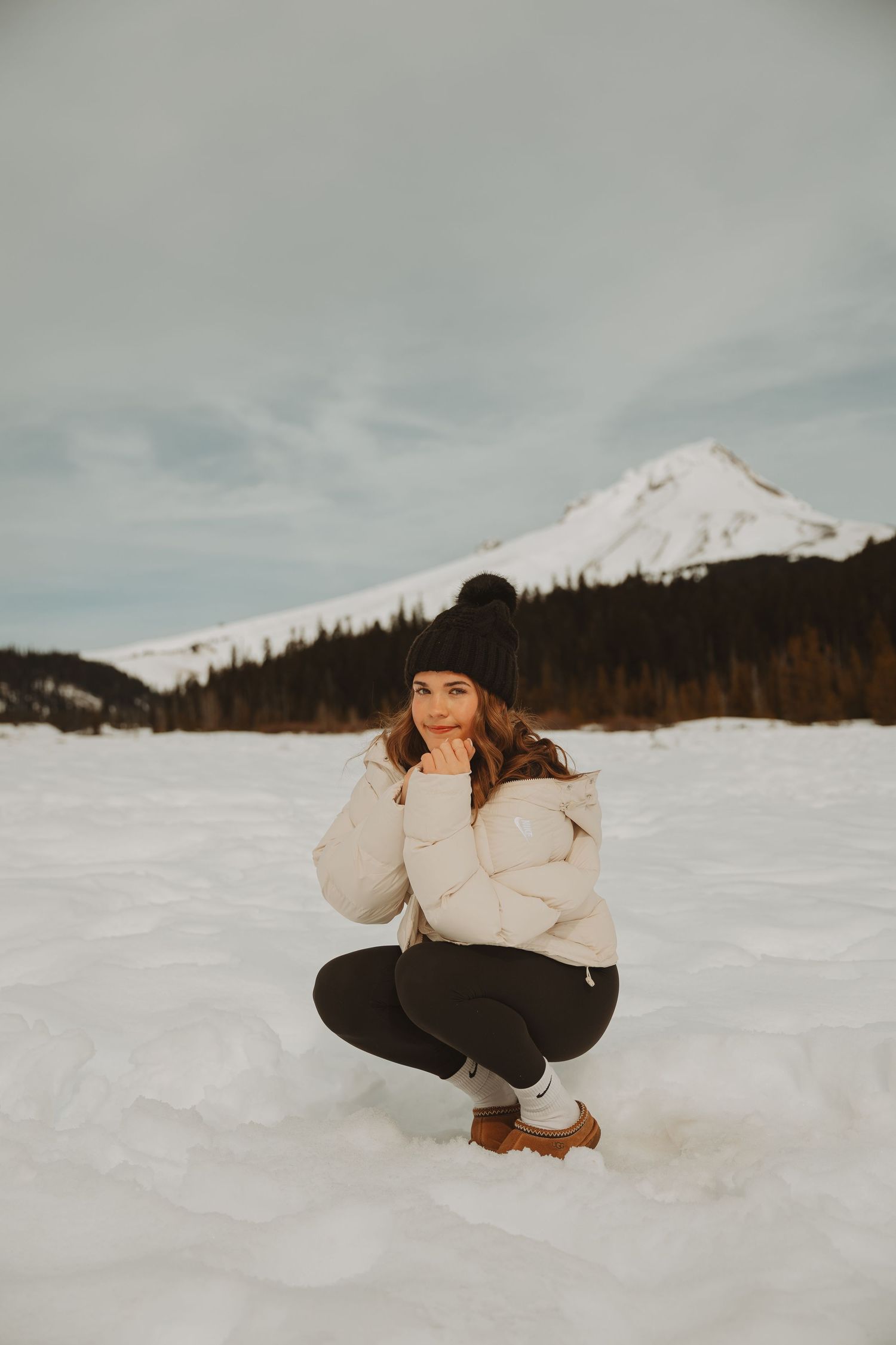 Young woman in white puffer jacket and black beanie crouches on snow with mountain in background.