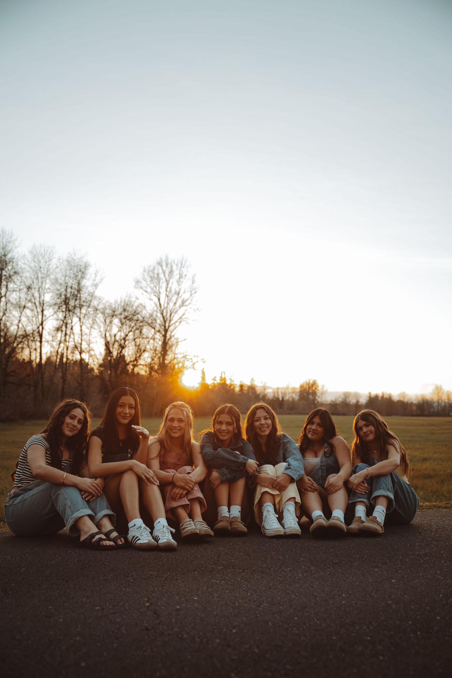 Seven young women sitting together on a road smiling at sunset with warm golden sky.