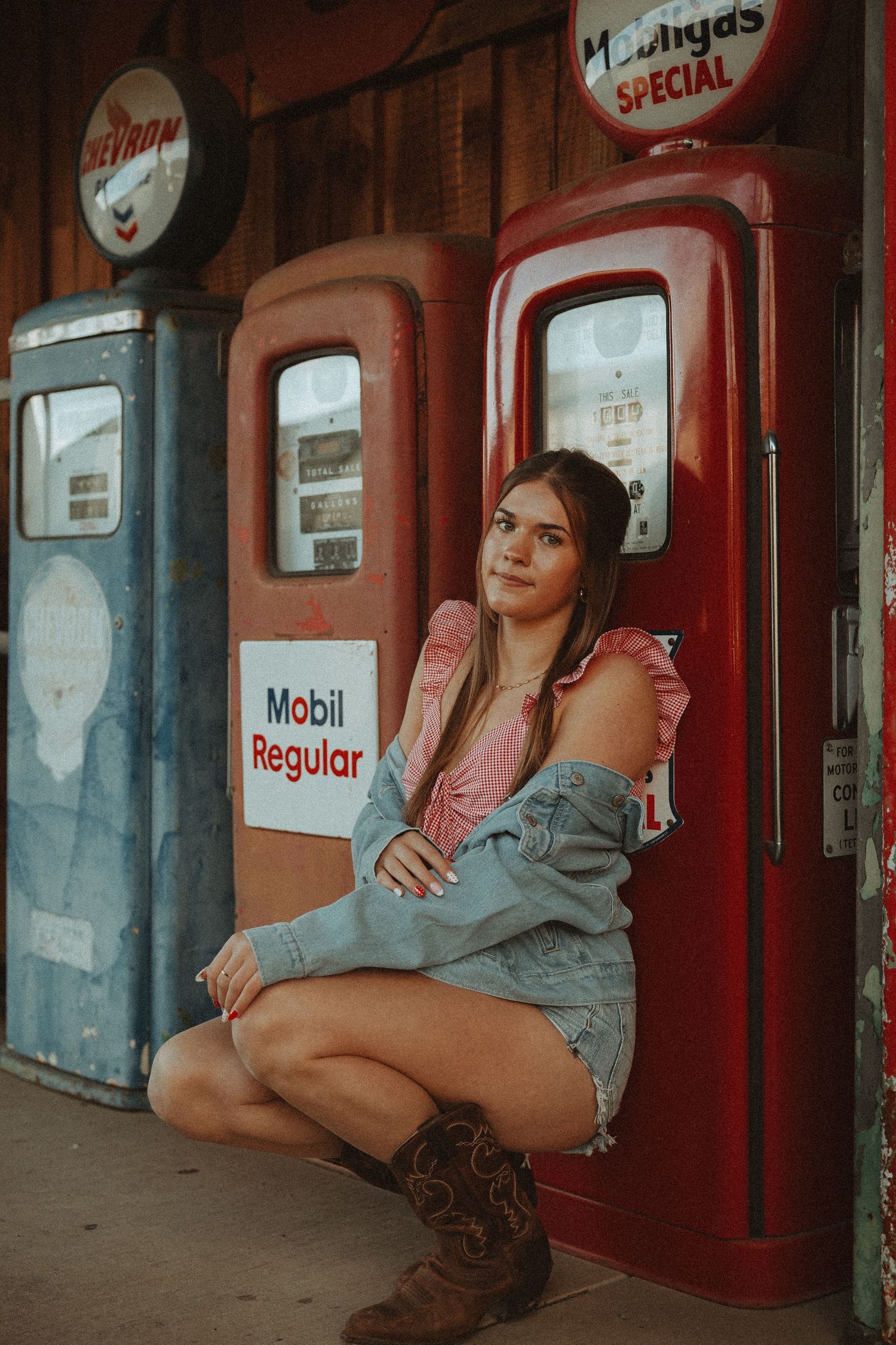 Young woman in denim jacket and cowboy boots posing by vintage Mobil Regular gas pumps at retro station.